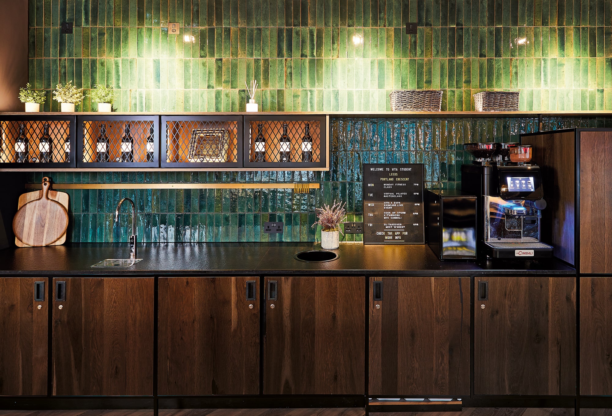 A modern kitchen corner with a dark wood countertop and cabinets. Green tiled backsplash, decorative plants, and a coffee machine sit alongside study materials. A metal grid shelf with bottles adds charm. Warm lighting creates an inviting atmosphere perfect for both cooking and studying.
