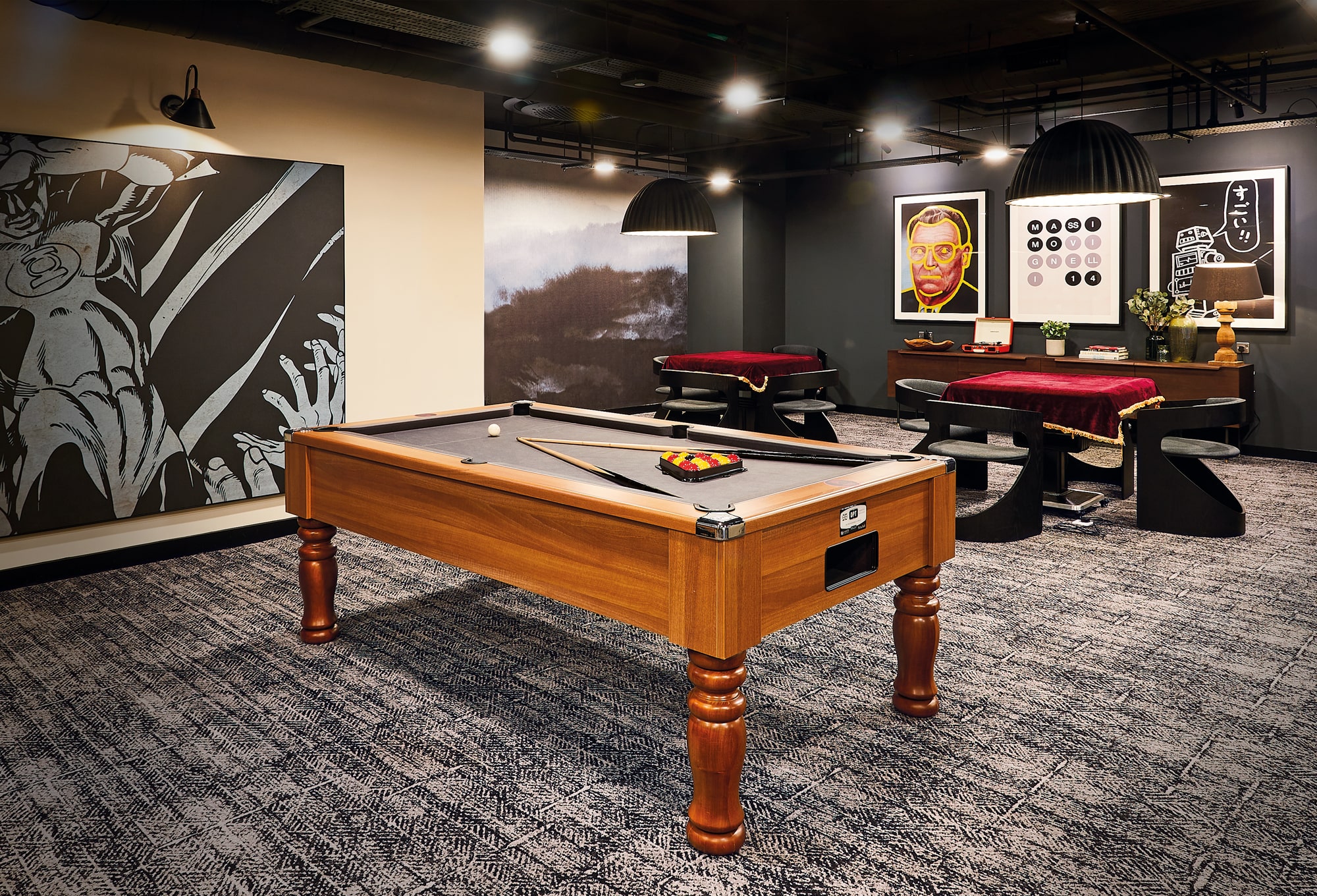 A stylish room with a pool table on a patterned carpet exudes a cozy, scholarly vibe. Artwork, including a black-and-white piece and an Indian-inspired colorful portrait, adorns the walls. Two red-clothed tables with unique chairs sit in the background under modern lighting.