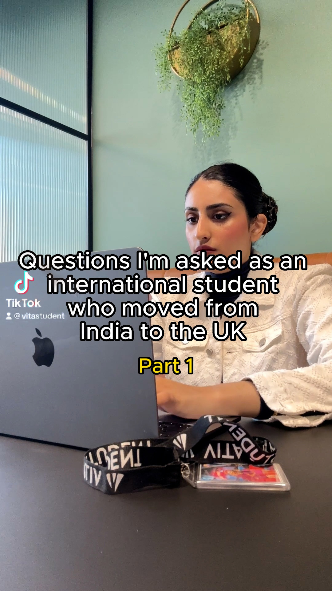 A woman sits at a table, typing on a laptop, with a phone and lanyard nearby. The text reads, "Questions I'm asked as an international student from India about studying in the UK, Part 1.