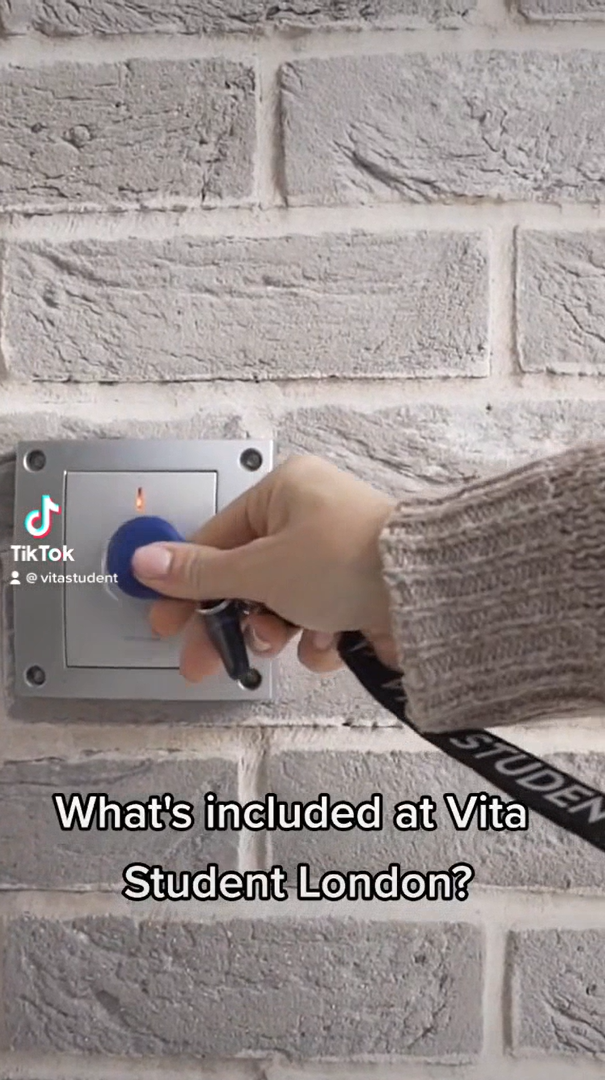 A hand in a beige sweater holds a blue key fob to a wall-mounted scanner on a light gray brick-patterned wall. Text reads, "What's included at Vita Student London?" A partially visible "STUDENT" lanyard hints at exciting opportunities for those studying abroad, whether from India or elsewhere.