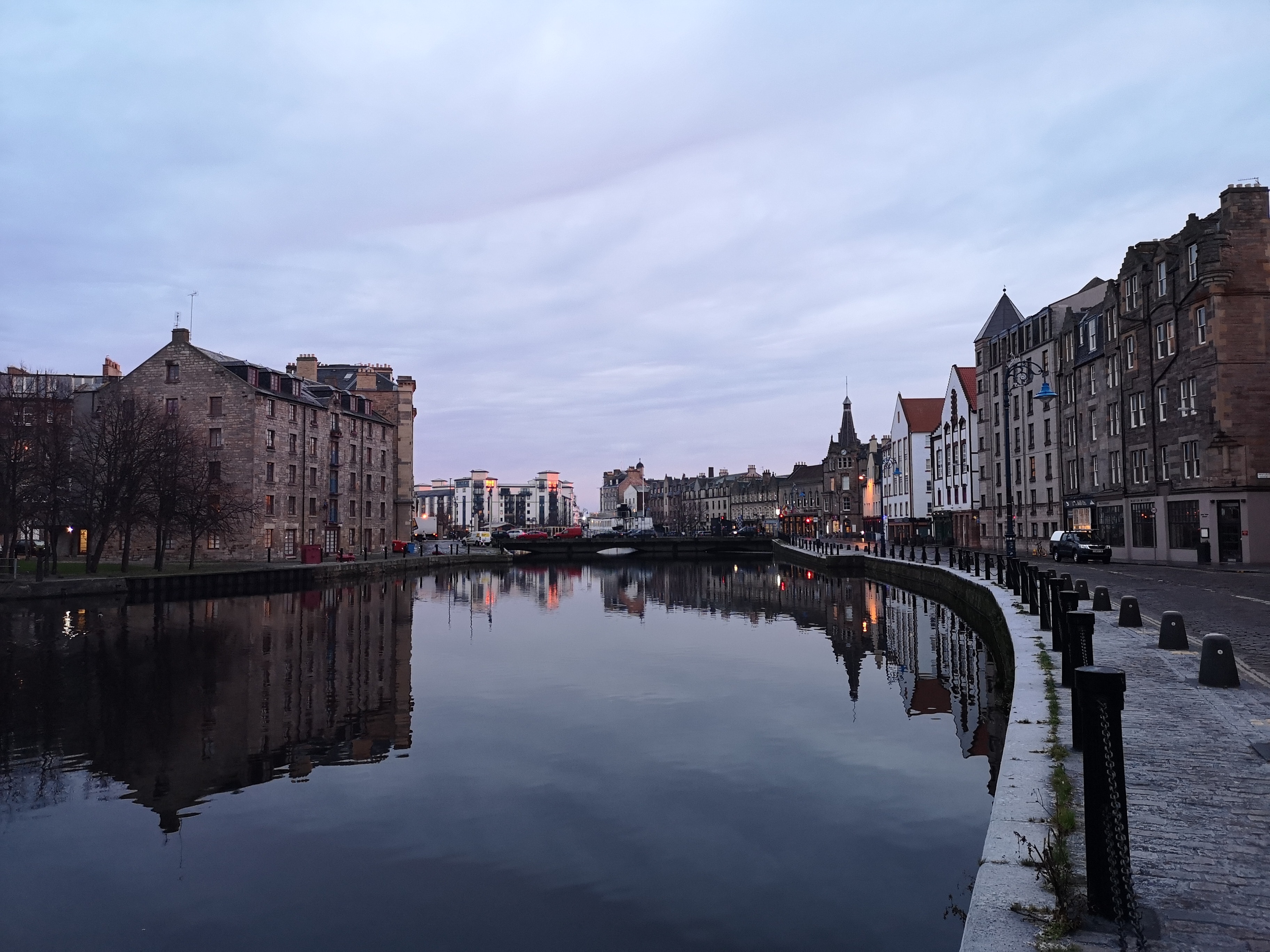A tranquil waterfront scene at dusk in Leith, reflecting historic buildings and a cloudy sky on the calm water. The street is lined with cobblestones and lampposts, creating a serene urban landscape reminiscent of Vita Student's contemporary charm.