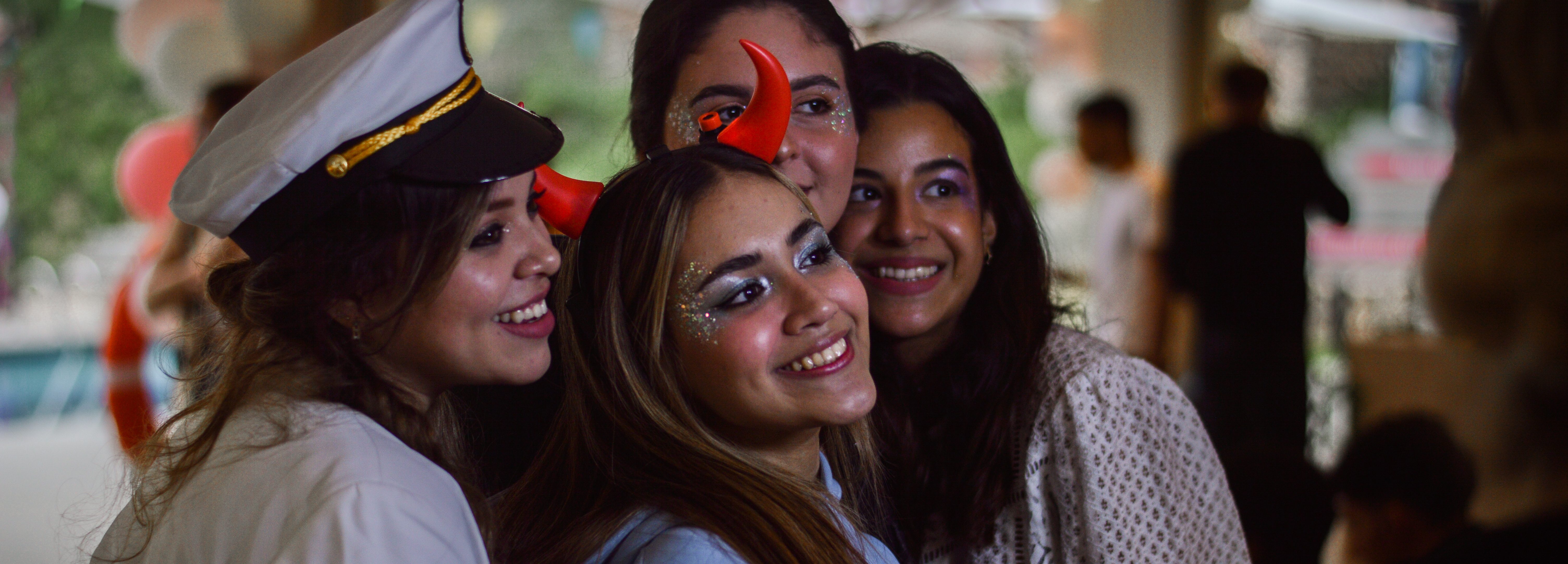 Four people smiling in a close group selfie. One wears a sailor hat, and two have red devil horns. Their glittery makeup sparkles with festive anticipation, expecting fun. The softly blurred background reveals others mingling next to vibrant decorations, capturing pure joy.