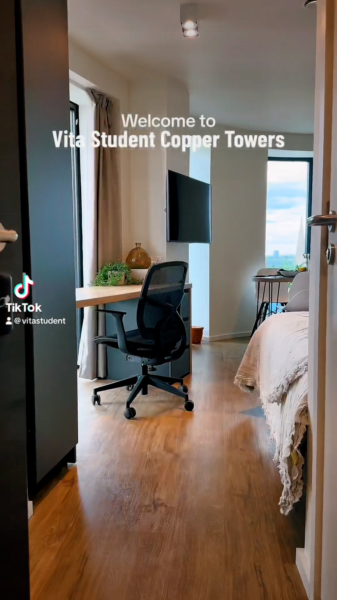 A cozy student apartment room with a wooden floor, ideal for studying. It features a bed with a beige duvet on the right and a black office chair at a wooden desk by a large window. A wall-mounted TV and potted plant are visible. Text reads "Welcome to Vita Student Copper Towers.