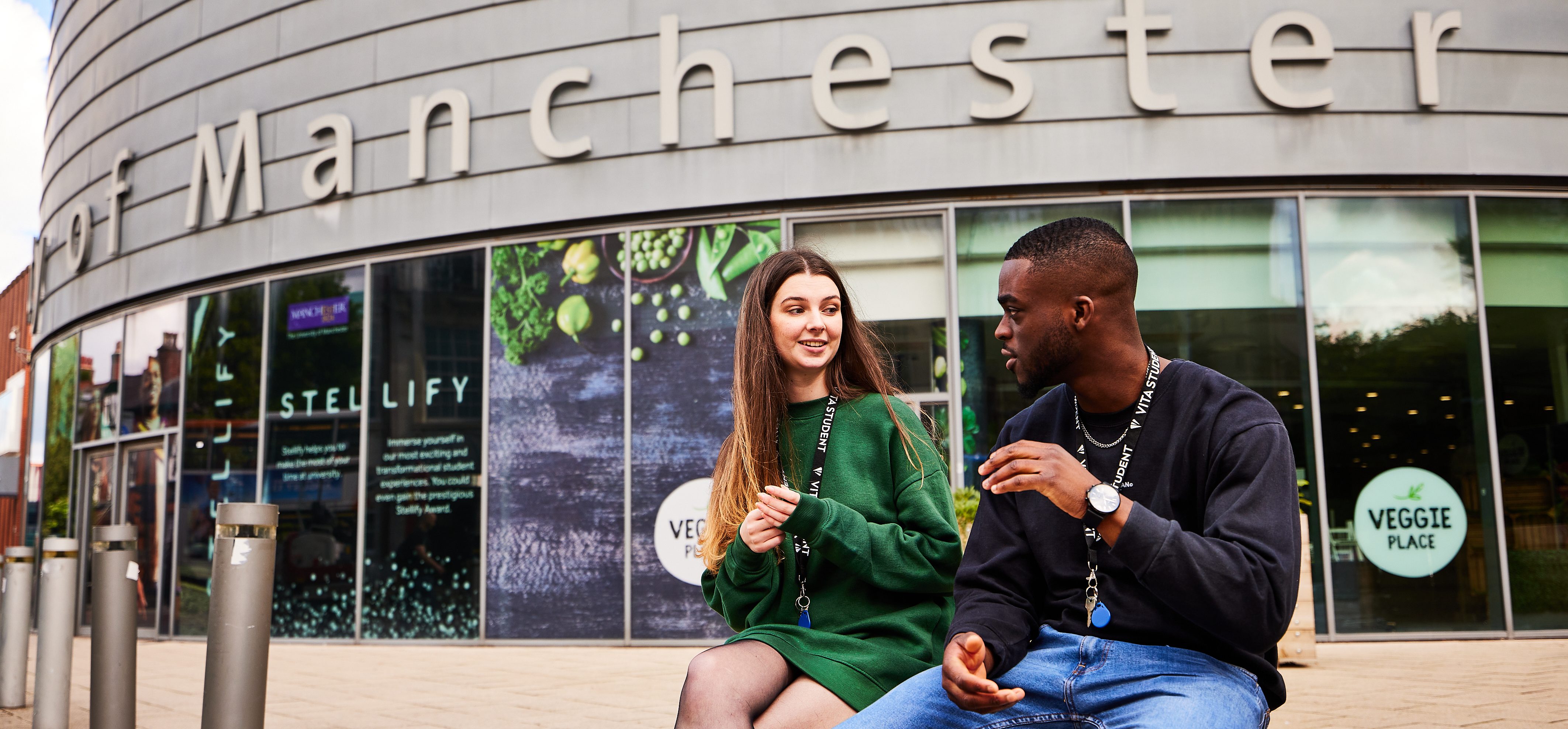 Two people sit chatting in front of a building adorned with large letters reading "Manchester." The woman, in a green sweater dress, and the man, in a black sweatshirt and jeans, seem to be discussing their choice of university degree. Behind them, signs for "VEGGIE PLACE" can be seen.