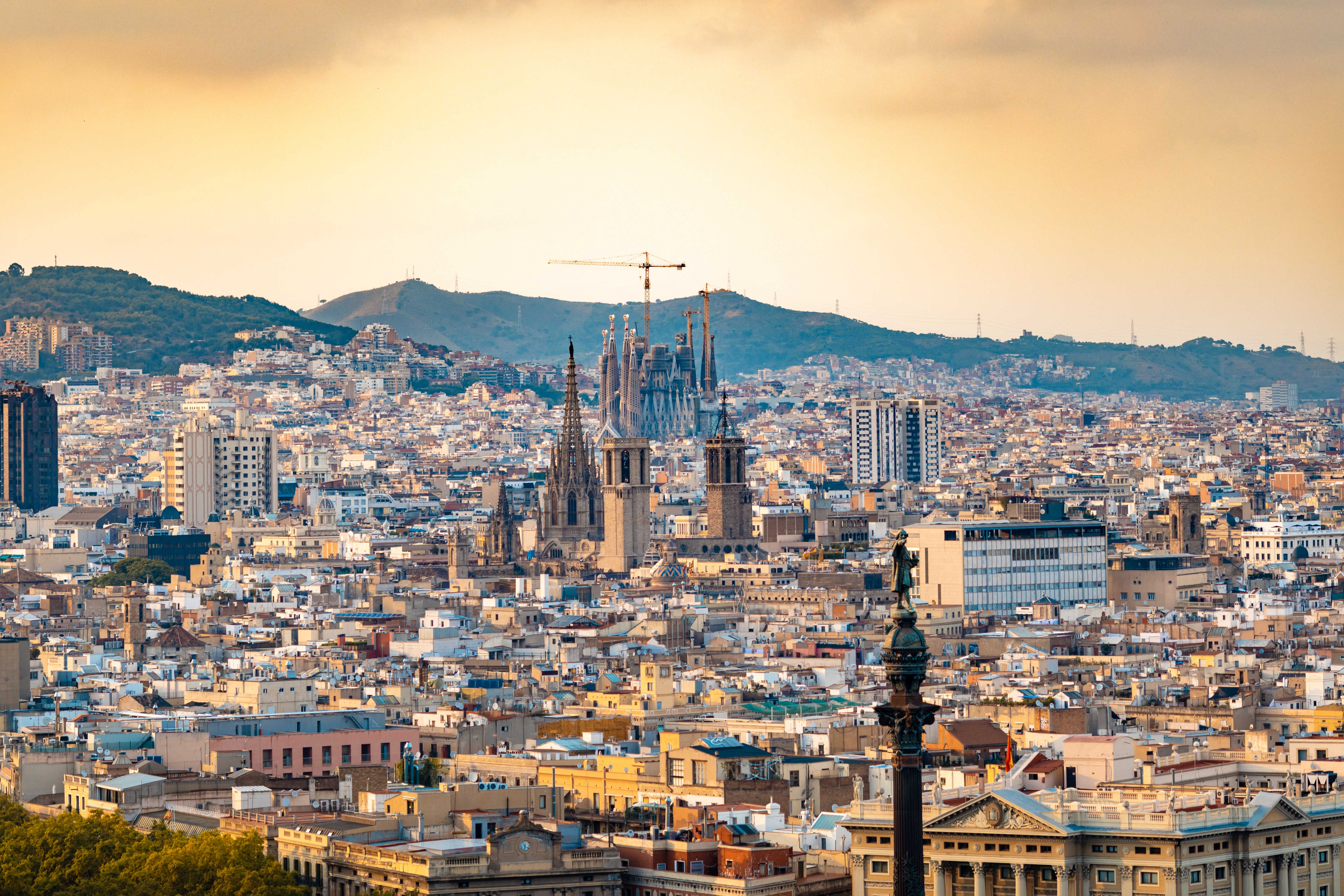 A panoramic view of Barcelona's cityscape at sunset features the iconic Sagrada Familia basilica prominently in the middle. Surrounding buildings are densely packed, while hills rise in the background under a golden sky—a perfect setting emphasizing razones para estudiar en Barcelona.