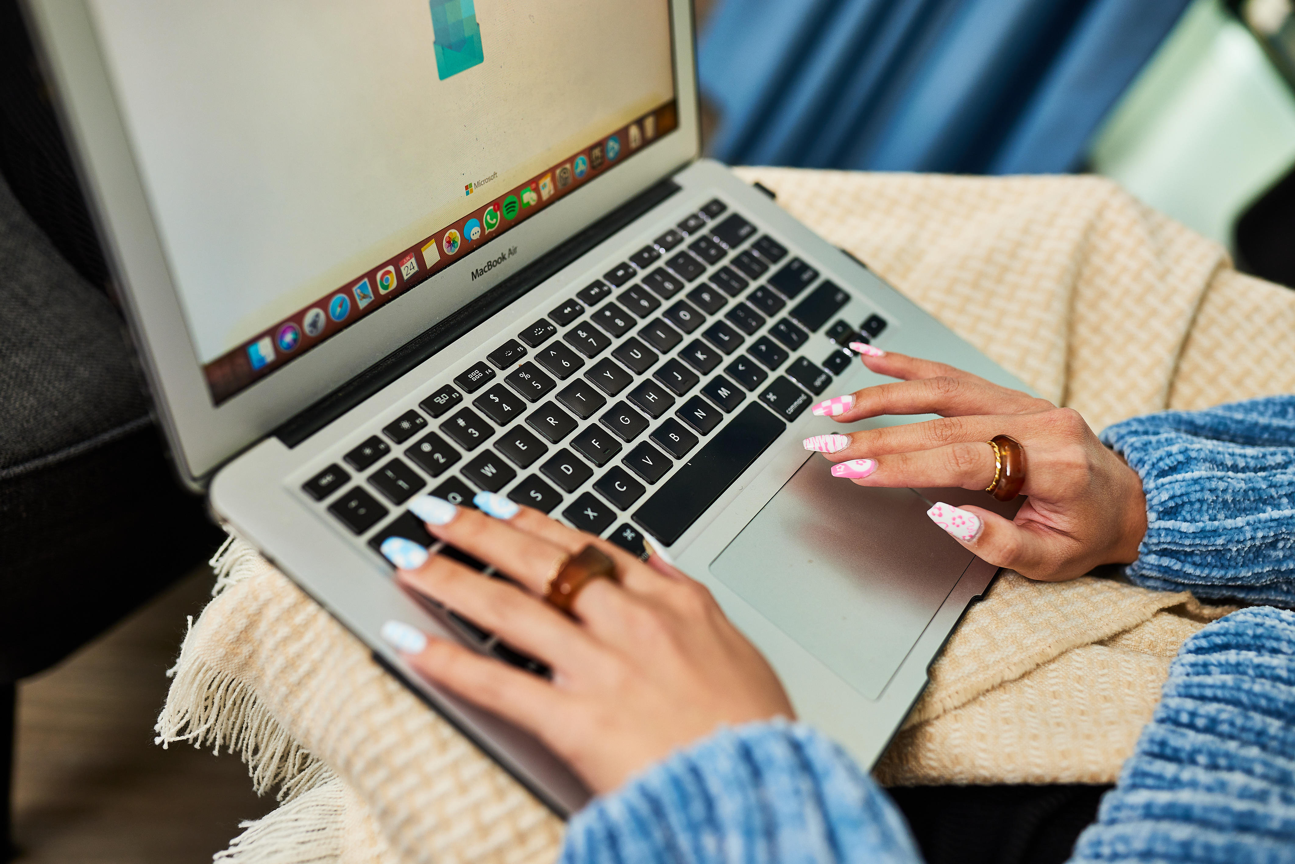 A person in a blue sweater types on a laptop resting on their lap, perhaps researching what to expect for the 2024 UK Results Day. A beige blanket partially covers their lap as vibrant, patterned nail art and multiple rings adorn their fingers. Behind them, a hint of a curtain completes the cozy scene.