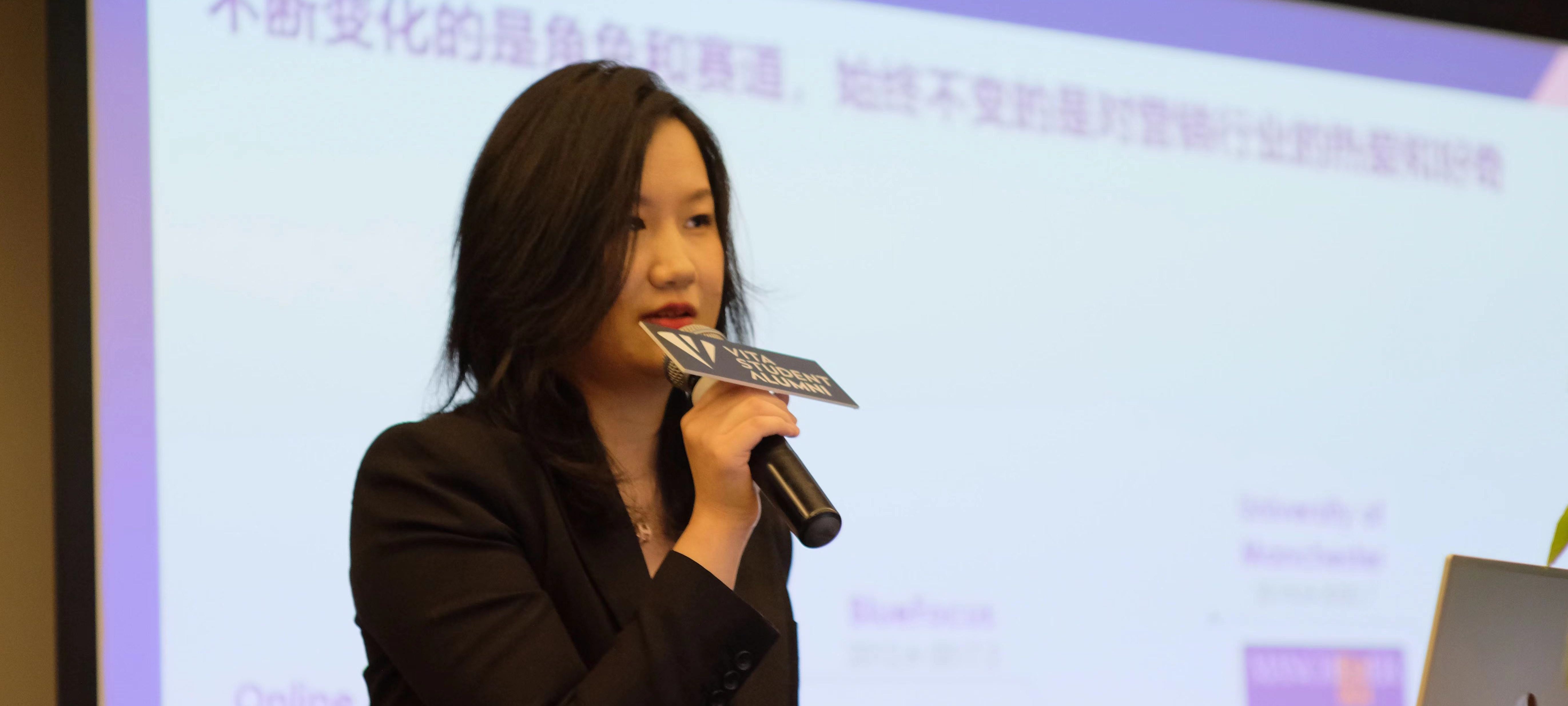 A woman in a black blazer speaks into a microphone at a presentation, embodying "Beyond Ambition." She holds a card with a logo. In the background, a blurred projection screen displays text in Chinese and English, setting the stage for Simple's story.