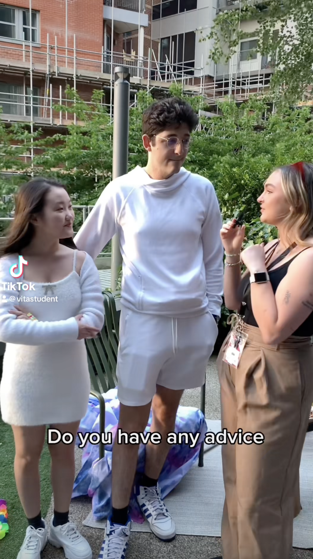 Three people are conversing outdoors. Two women stand on either side of a man wearing white. One woman holds a microphone. They are surrounded by lush greenery with a building under construction in the background. The text on screen reads, "Beyond Ambition: Do you have any advice?.