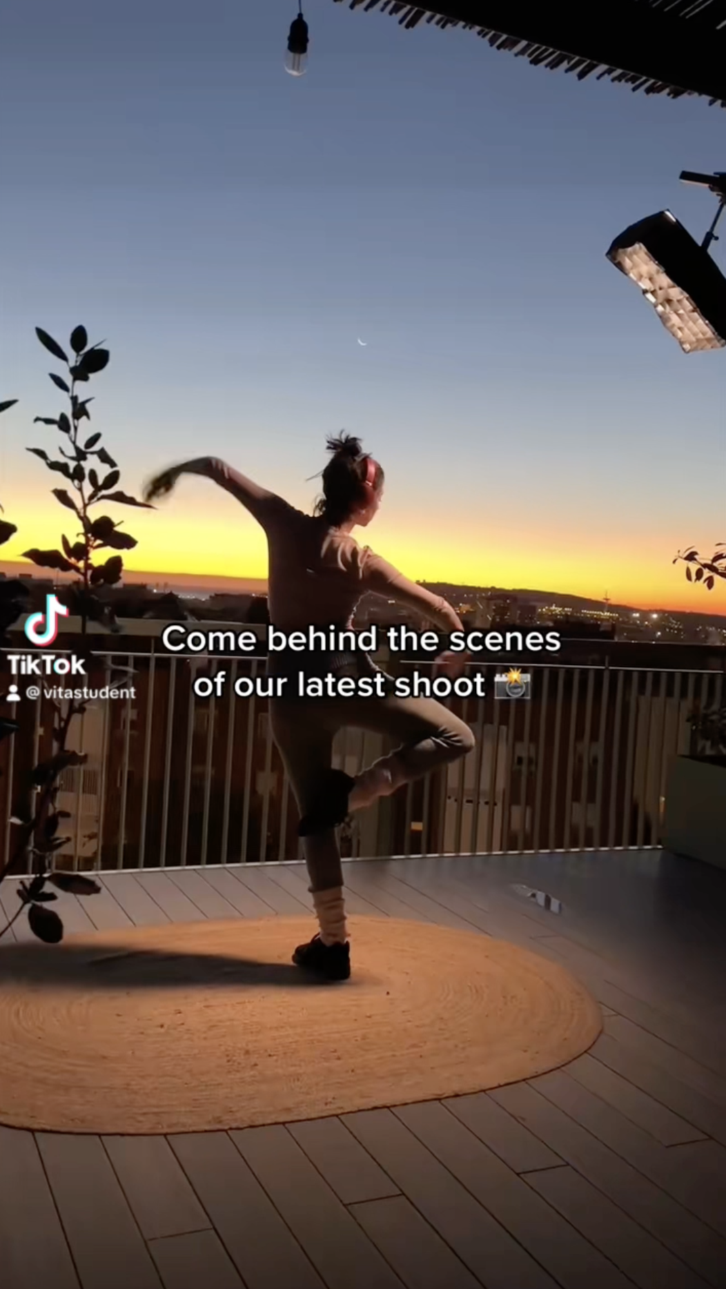 A person is posing gracefully on one leg against a balcony railing at sunset, embodying a sense of beyond ambition. A crescent moon graces the sky, plants surround the circular mat beneath them, and city lights shimmer in the background.