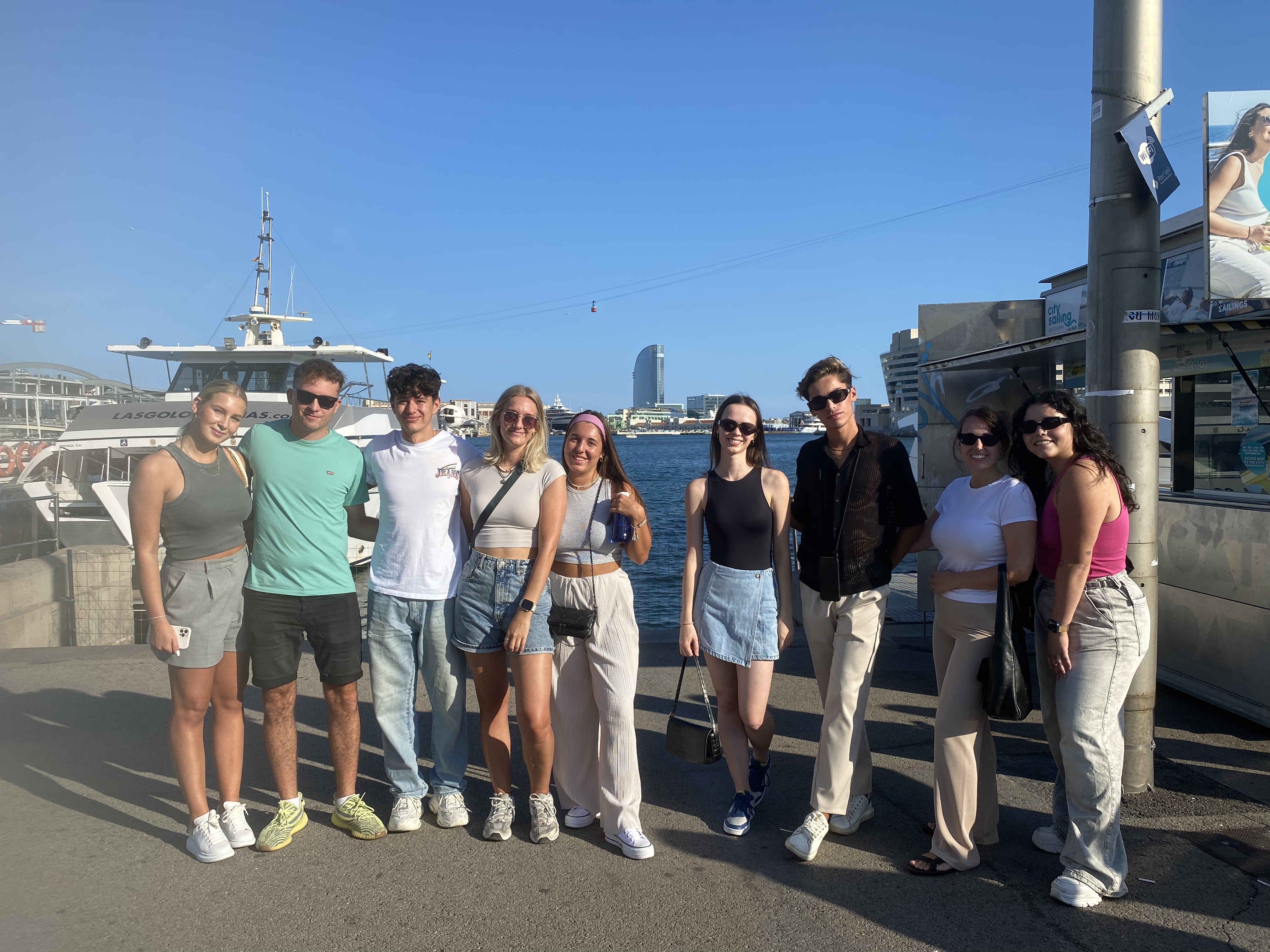 A group of nine people, possibly students, stands smiling near a dock in sunny Barcelona, with a boat and the ocean behind them. Casually dressed, they're enjoying the day with a city skyline visible in the distance—a snapshot from their guía definitiva for internacionales exploring new horizons.