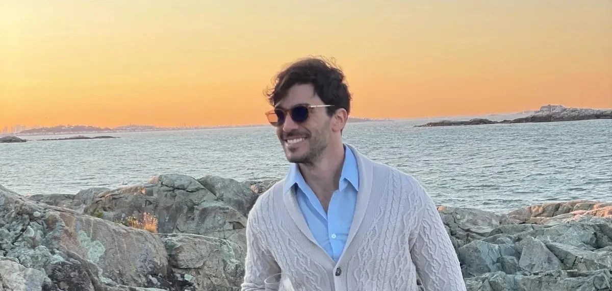A person wearing sunglasses and a light sweater smiles while holding a drink. They stand on rocky terrain near a body of water, embodying the essence of Juan's story. The sunset casts an orange glow over the scene, highlighting a moment that transcends ambition.