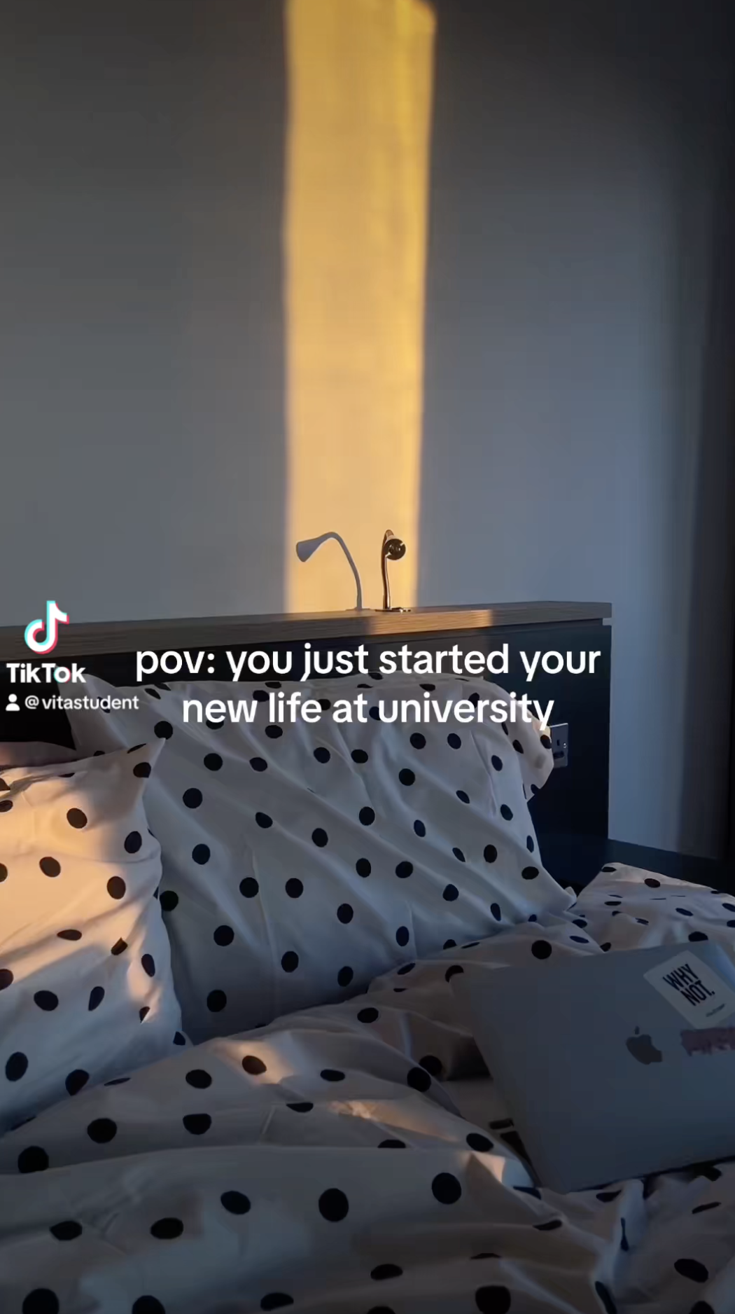 Sunlight casts a warm glow on a neatly made bed with polka dot pillows and a wooden headboard. An open laptop sits on the bed, embodying ambition and personal growth as the text reads, "pov: you just started your new life at university.