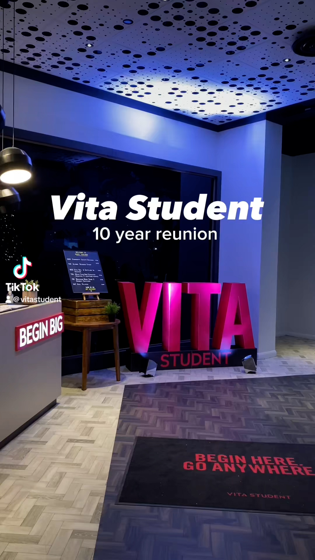 A modern room with a bright pink "VITA" sign and a poster for a "Vita Student 10 year reunion." The floor mat reads "BEGIN HERE GO ANYWHERE." The ambiance, illuminated by spotlights and a blue ceiling, inspires beyond ambition.