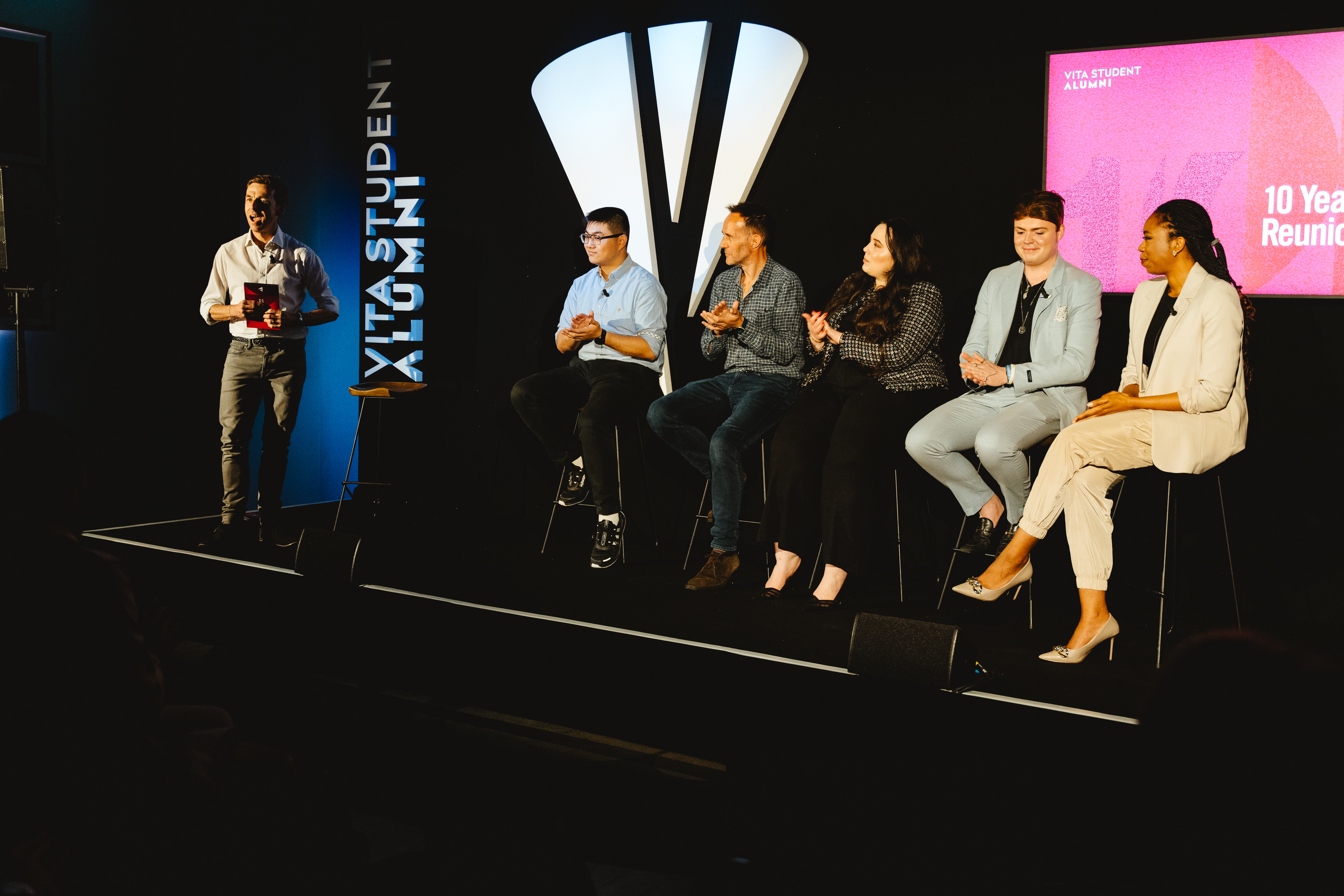A group of five people sit on stools on a stage, participating in a 10-year reunion event. They are dressed in business casual attire. Another person stands to the side, appearing to introduce or present them to an audience.
