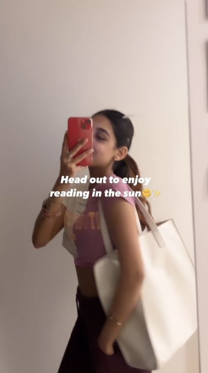 A person wearing a pink tank top and holding a white bag takes a mirror selfie with a red phone. Overlaid text reads: “Head out to enjoy reading in the sun, whether you're studying or just relaxing in vibrant India.”.