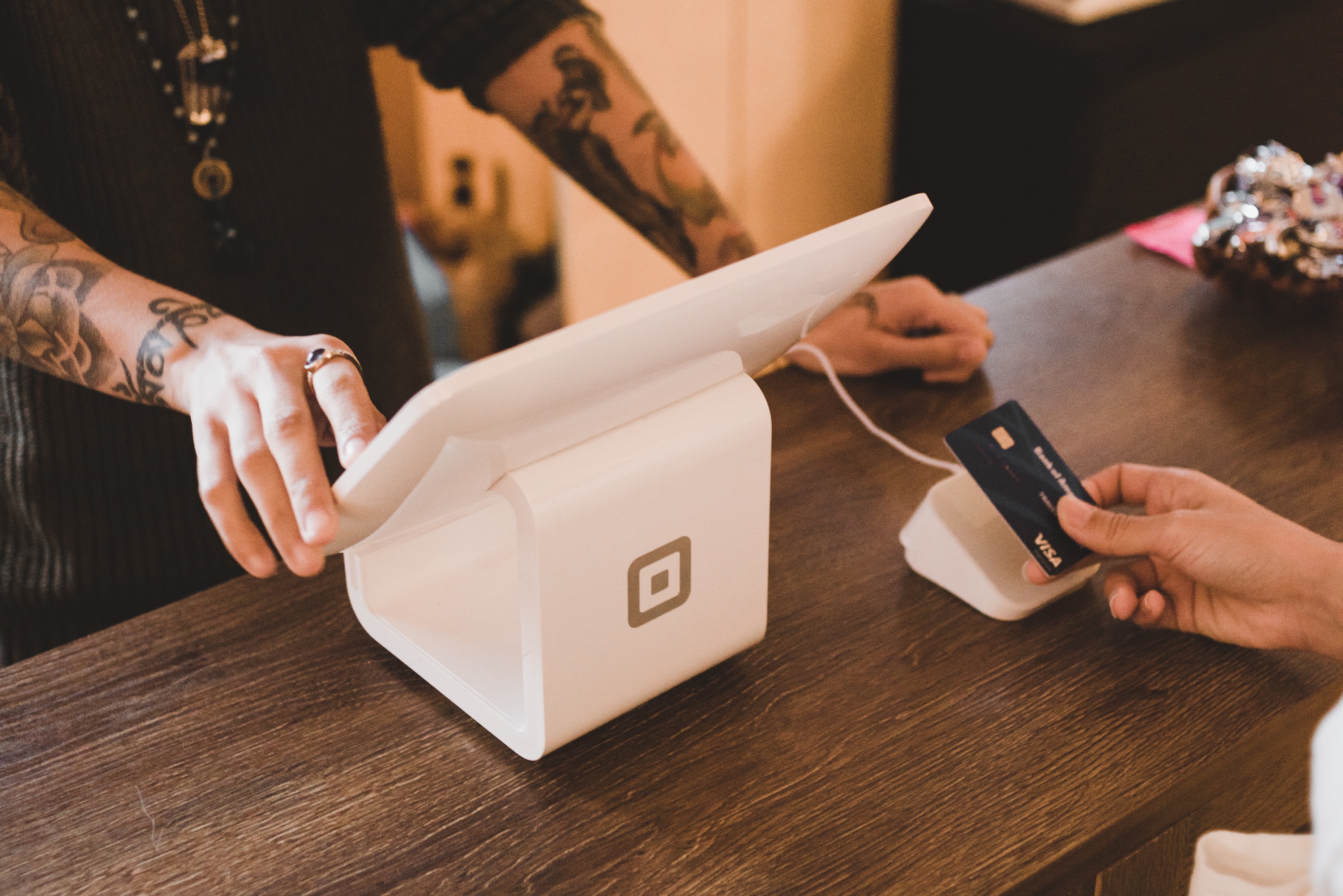 A person is making a payment using a blue credit card at a point-of-sale terminal on a wooden counter. An international student with tattooed arms is operating the touchscreen device attached to the terminal, perhaps managing their UK bank account transaction efficiently.