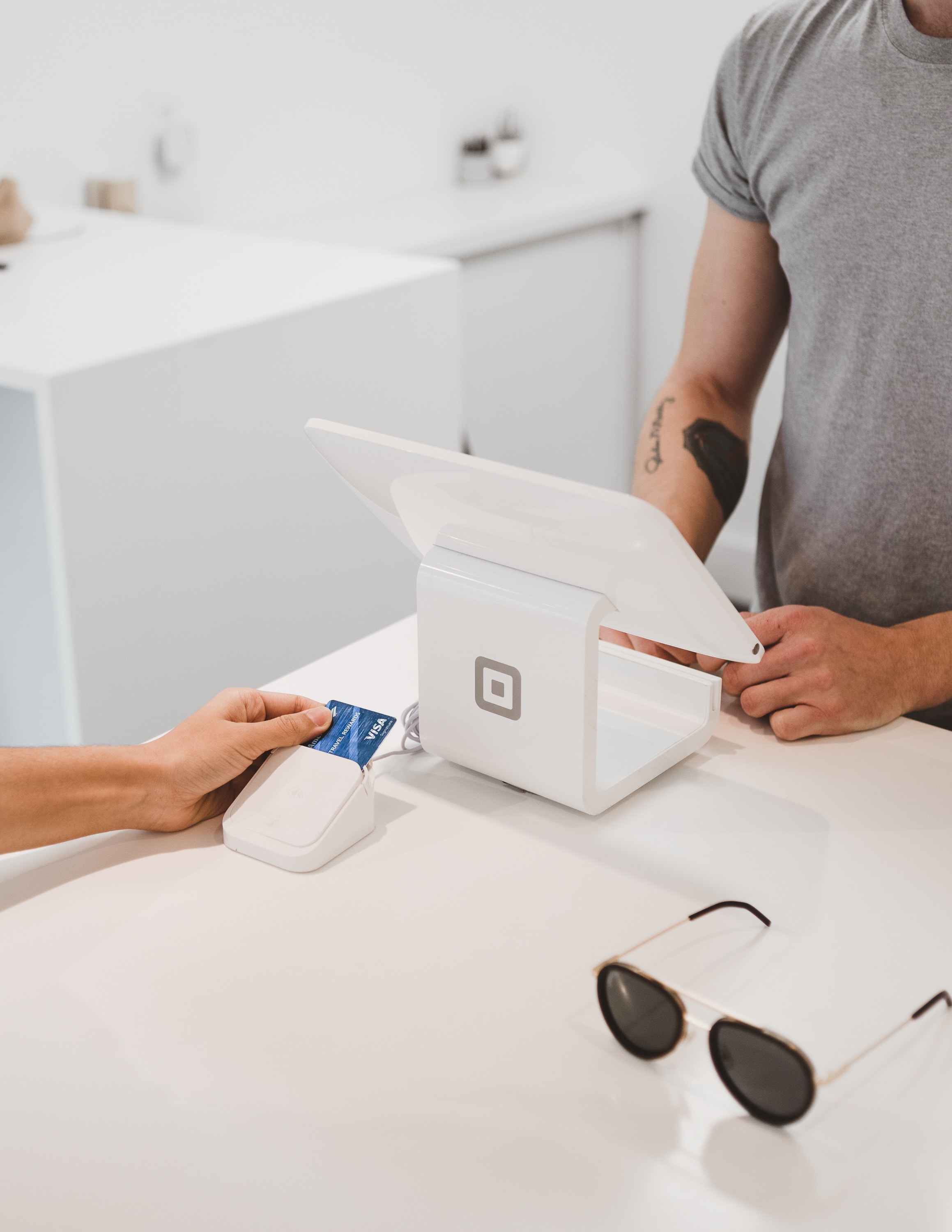 A person in a gray shirt stands at a white point-of-sale counter, holding a debit or credit card near a card reader. Sunglasses rest on the counter next to the reader. This modern payment transaction feels seamless, much like managing one's cuenta bancaria while studying as an estudiante internacional in España.
