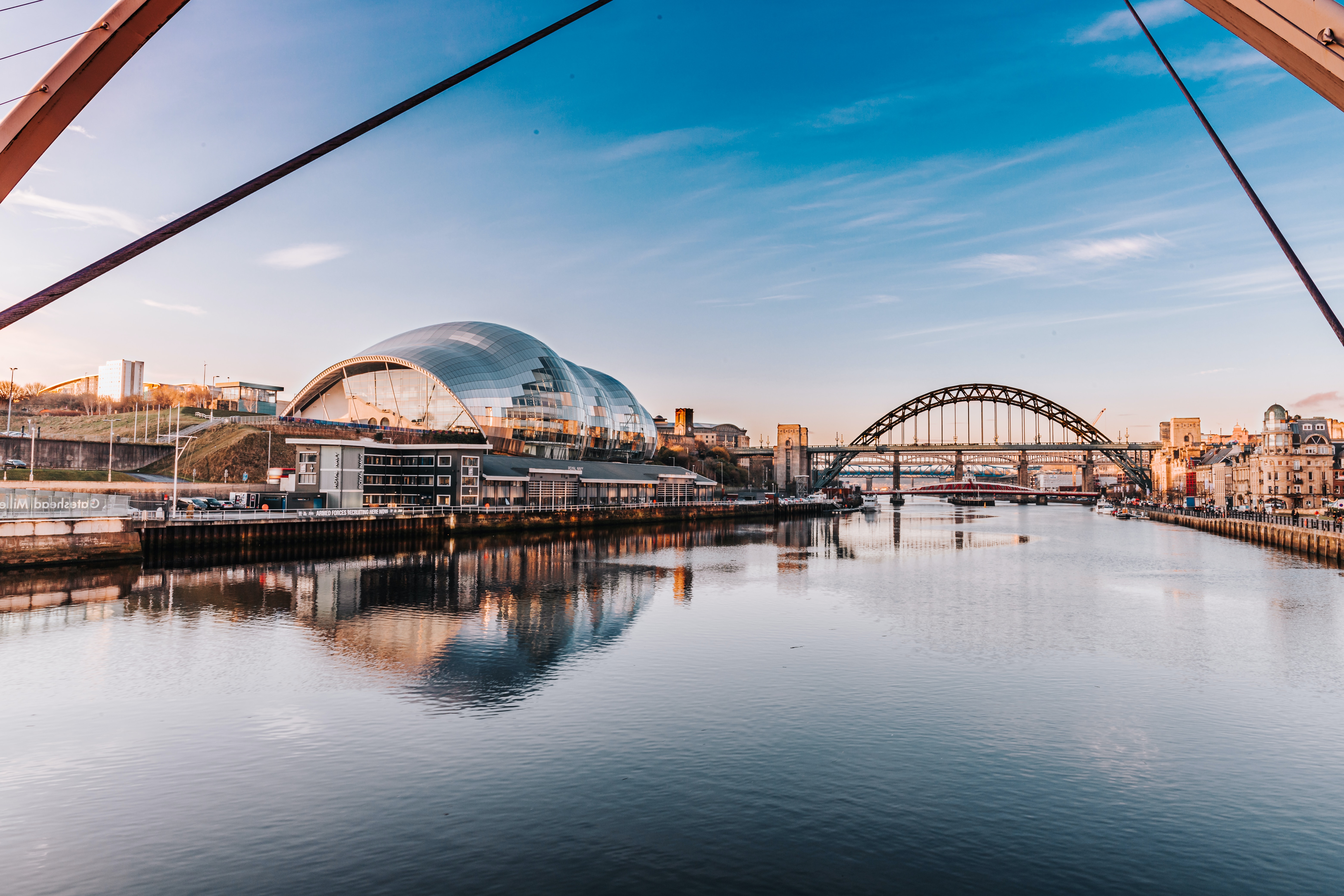 A scenic view of the River Tyne with modern, arched bridges spanning the water greets you. The skyline boasts a futuristic, curved building with glass panels reflecting the evening light—perfect for making the most of a parents' visit while exploring Newcastle Uni and its surrounding charm.
