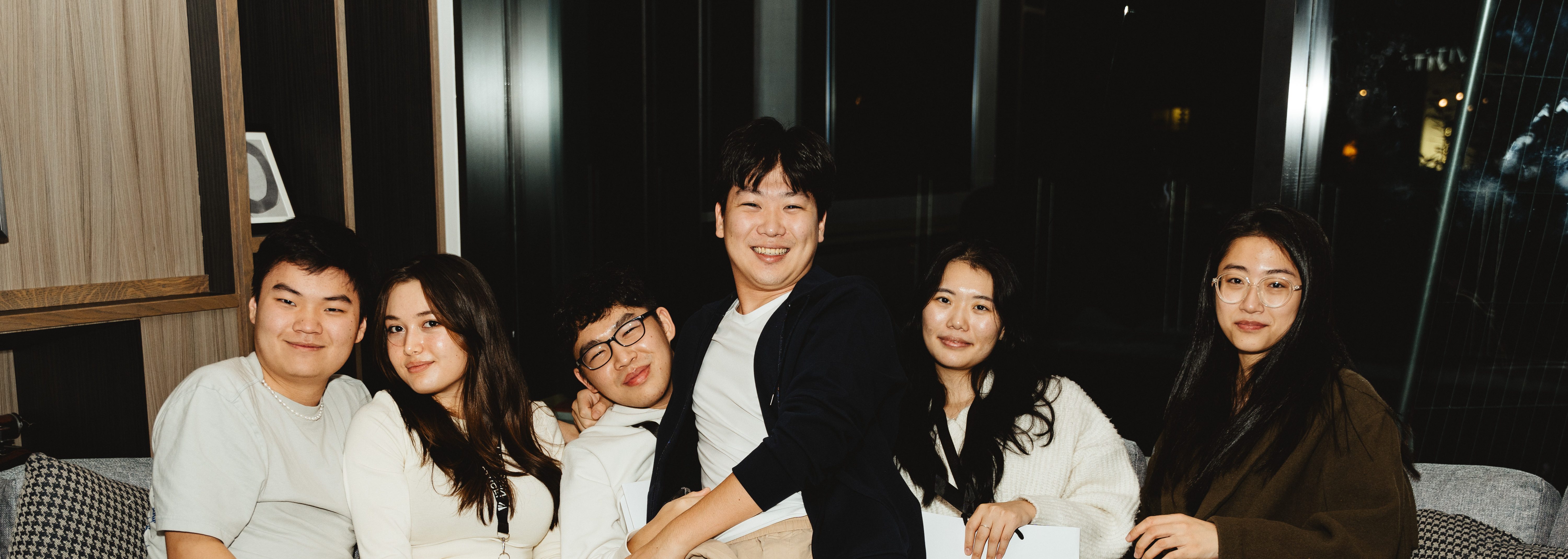 A group of six friends, celebrating their 10-year reunion, sits closely together on a couch, smiling warmly at the camera. In the cozy indoor setting with dim lighting, shelves and windows are visible in the background.