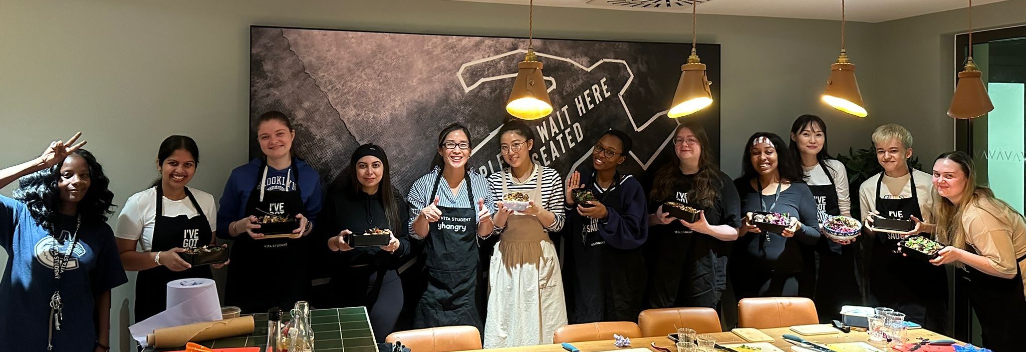 A group of eleven people stand in a line, each holding a decorated piece of clothing. They are in a room with pendant lights and a chalkboard wall filled with student recipes. Some wear aprons and smile at the camera, while one person makes a peace sign.