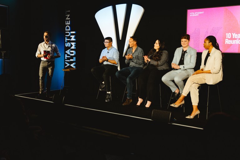 A group of five people sits on stools on a stage under a spotlight, engaging in a panel discussion. They are smiling and clapping. A man stands to the left holding a microphone. A screen in the background reads "10 Year Reunion.