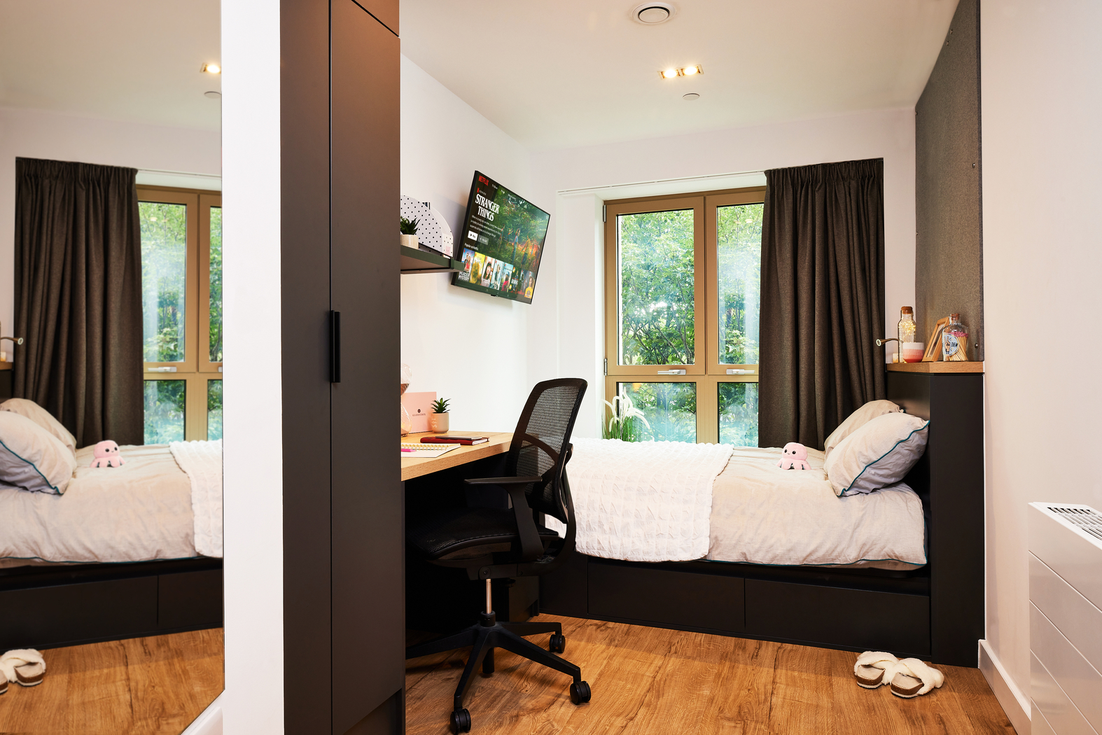 A cozy bedroom with a single bed placed against a wall, featuring white bedding and plush toys. There's a desk with a chair beneath a mounted TV, a plant, framed photos, and a large window with curtains overlooking greenery.