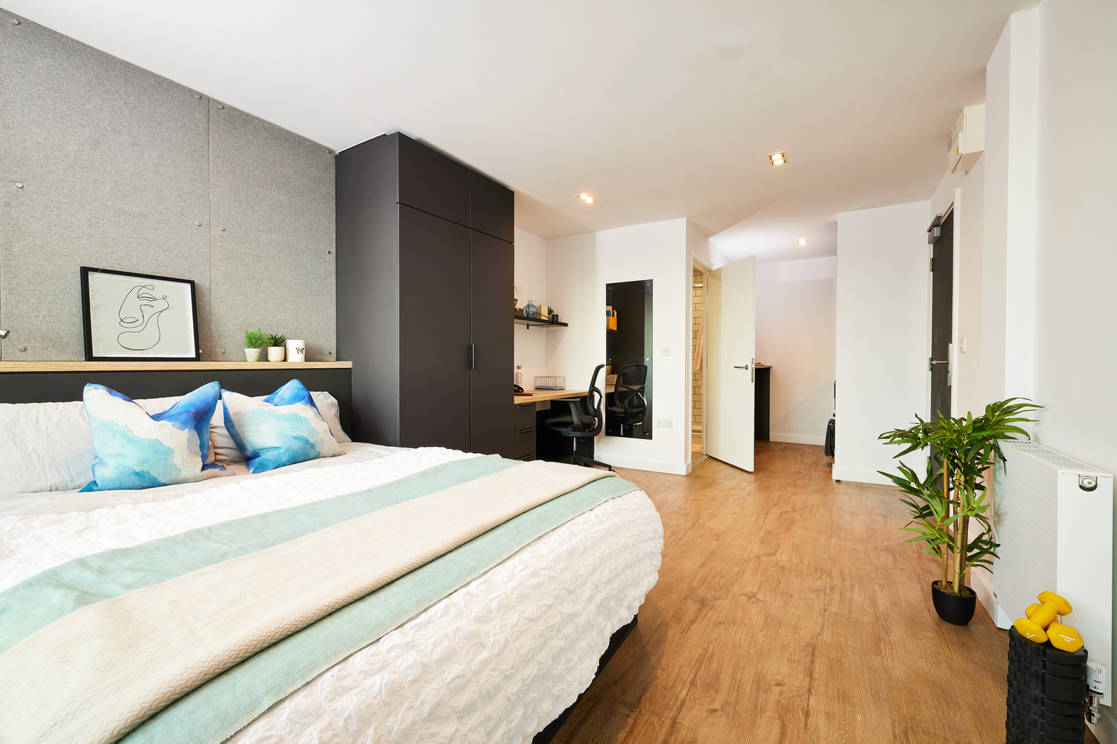 Modern bedroom with a large bed featuring blue and white pillows and a light green throw. The room has wooden flooring, a black wardrobe, a desk with a chair, indoor plants, and soft ambient lighting. A small framed artwork is on the wall.