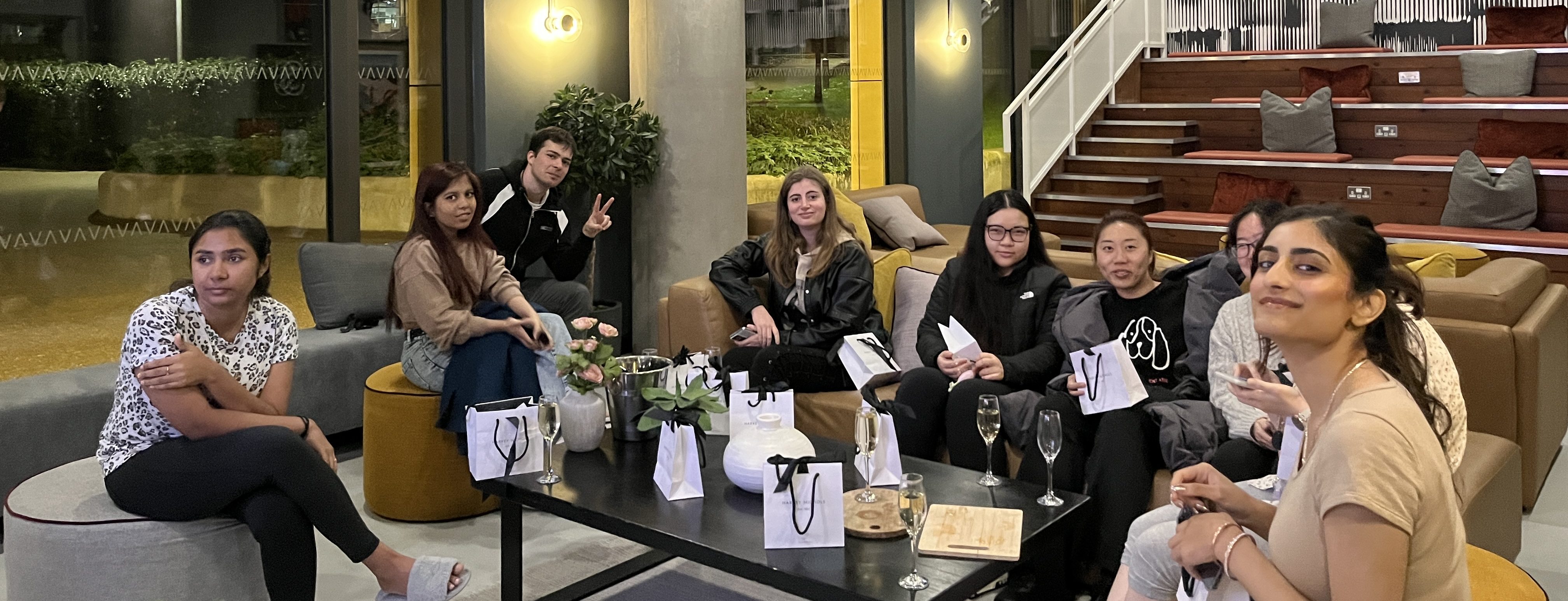 A group of people sitting together in a cozy indoor setting with modern decor, reminiscent of a chill spot to study at uni. They appear relaxed, some holding gift bags and drinks. A staircase and greenery are visible in the background.