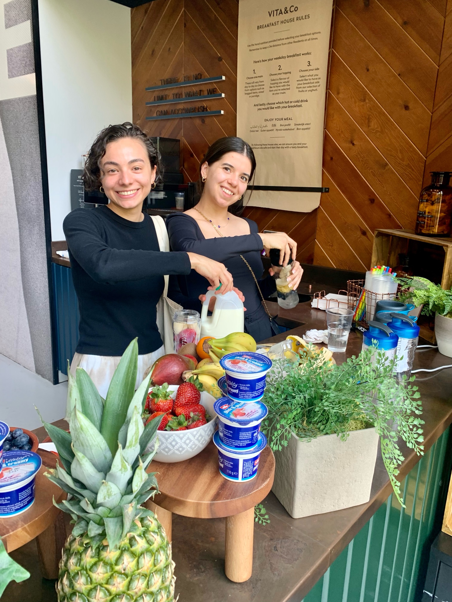 Two women are smiling while preparing smoothies at a wooden counter. The counter has fruit like strawberries, bananas, and a pineapple, along with yogurt containers and leafy plants. They are in a cozy space with a menu visible in the background.