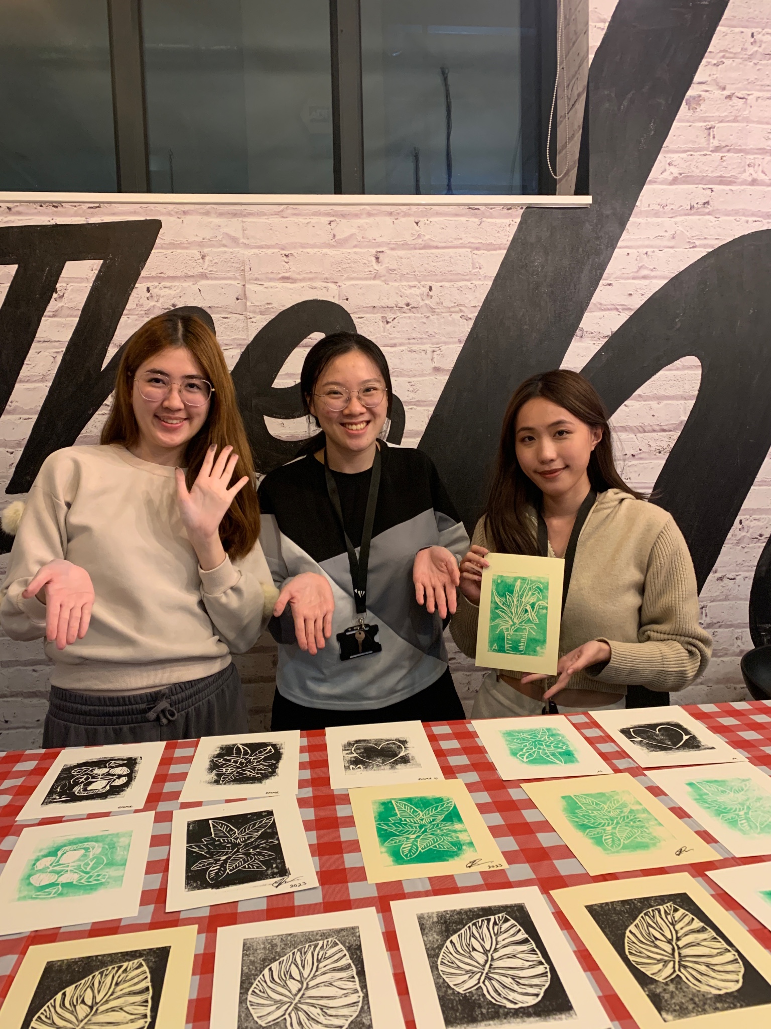 Three people smile at the camera while standing behind a table covered with leaf-themed artwork. The person on the right holds up one of the pieces. A brick wall with large lettering is in the background.
