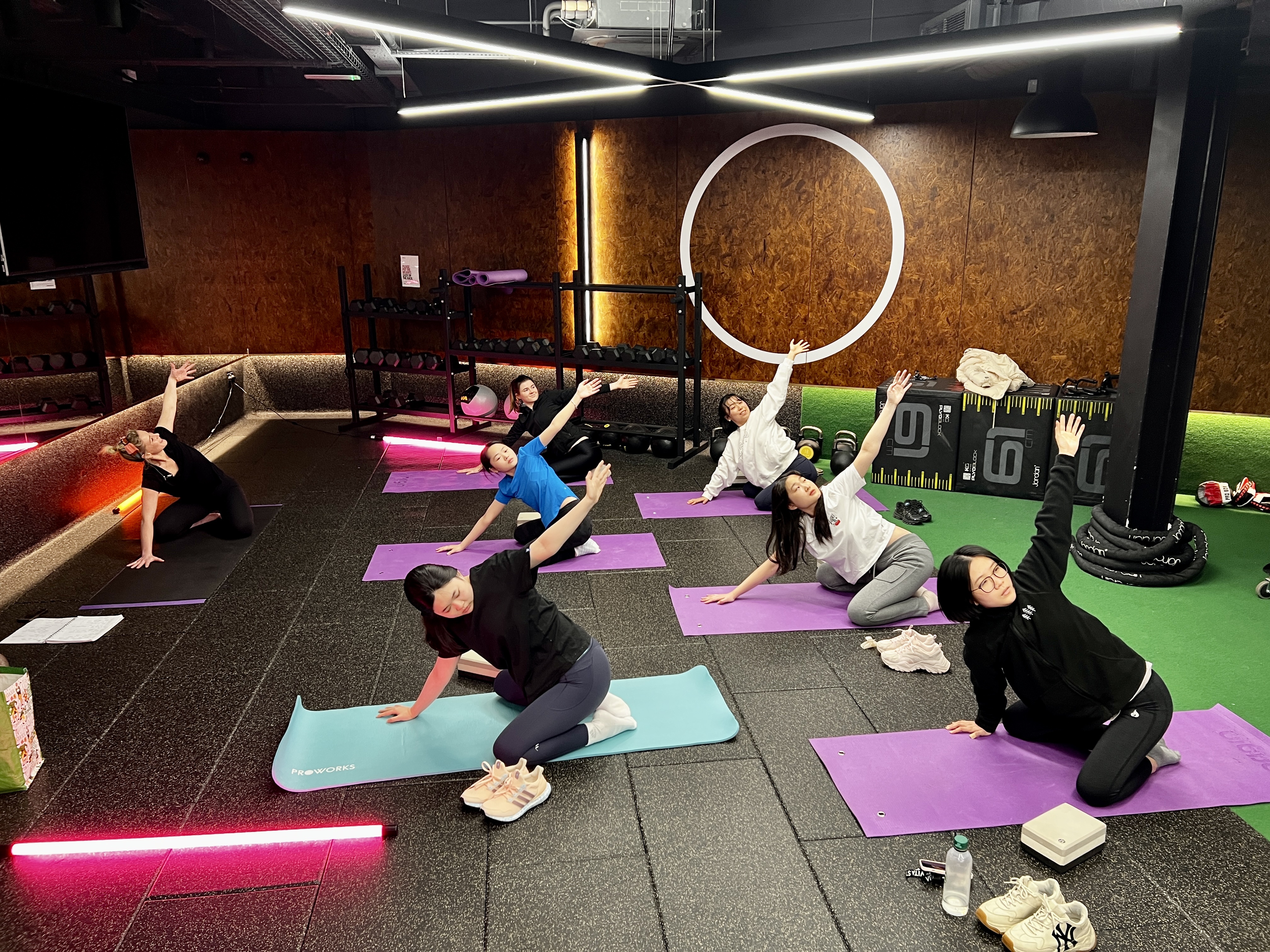 A group of people in workout attire are doing side stretches on yoga mats in a gym. They appear engaged and relaxed, with gym equipment visible in the background. The lighting is dim with a modern, industrial design.