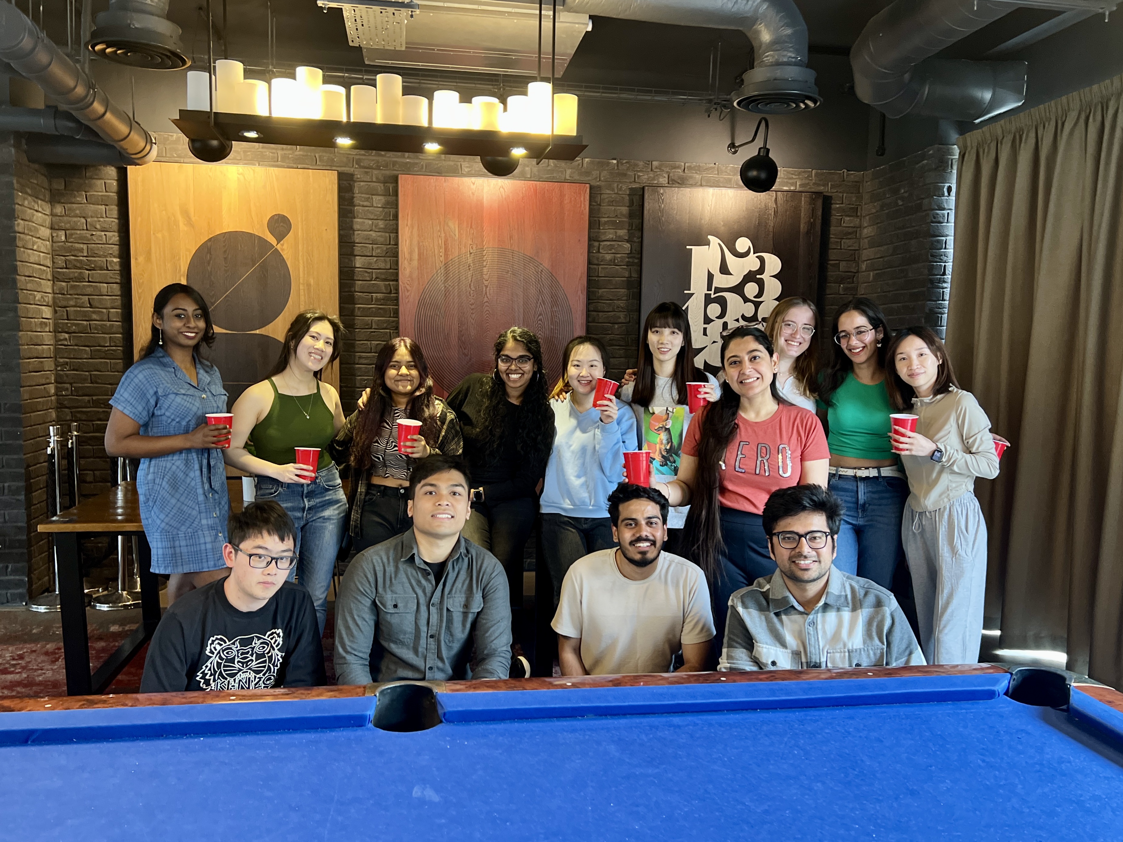 A group of people gathered in a room with a pool table, holding red cups. Some are sitting on the pool table, others standing behind it. The room has dark brick walls and framed artwork. The atmosphere appears lively and relaxed.