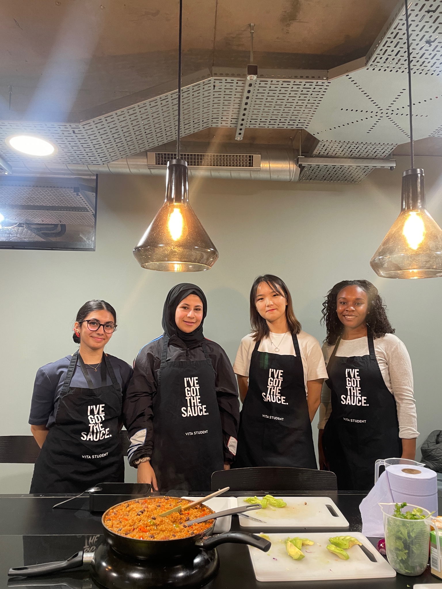 Four people stand together wearing black aprons with "I've Got the Sauce" printed on them. They are in a kitchen setting with a stove, two pendant lights above, and a prepared dish in a pan on the counter.