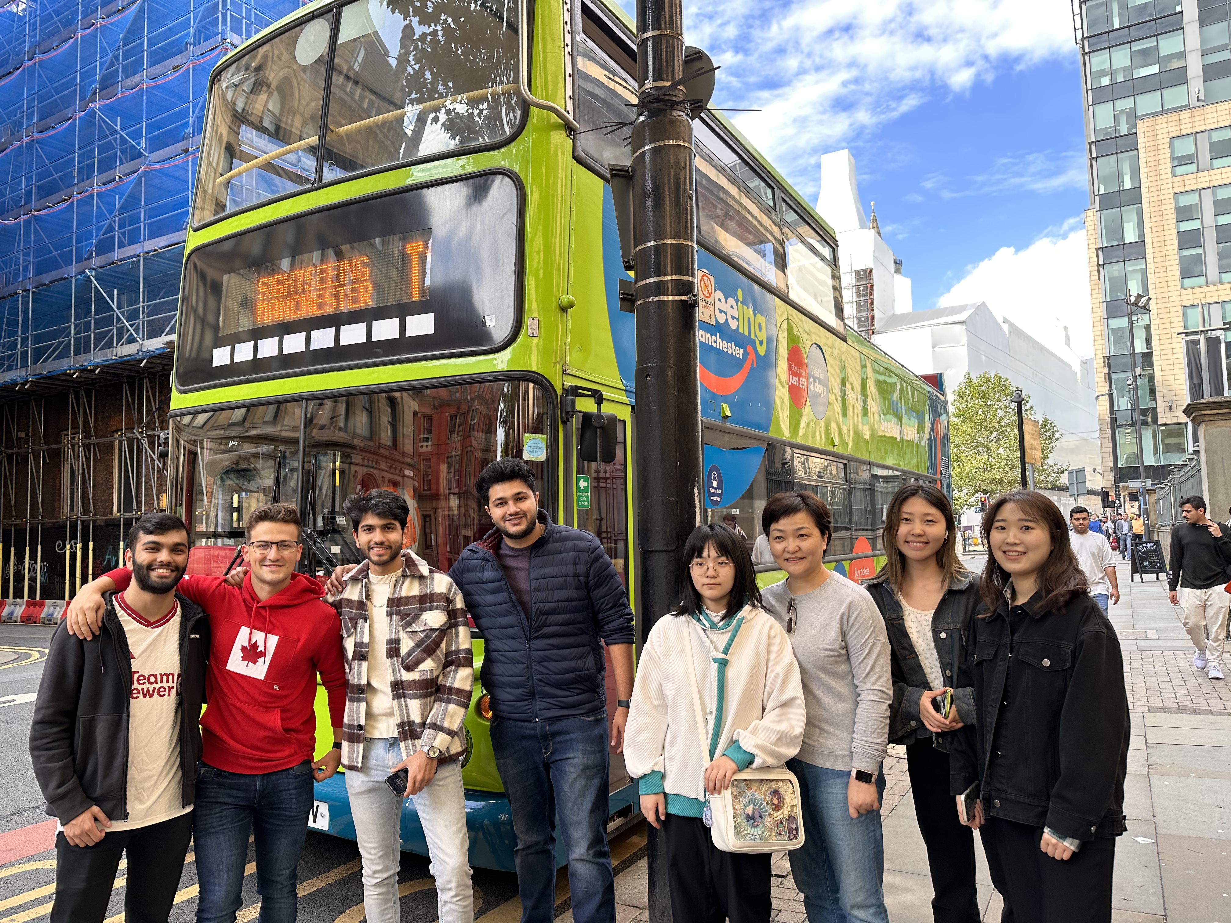 A group of eight people stands smiling in front of a green double-decker bus on a city street. The bus displays a route number and "Manchester." Buildings and a blue sky are visible in the background.