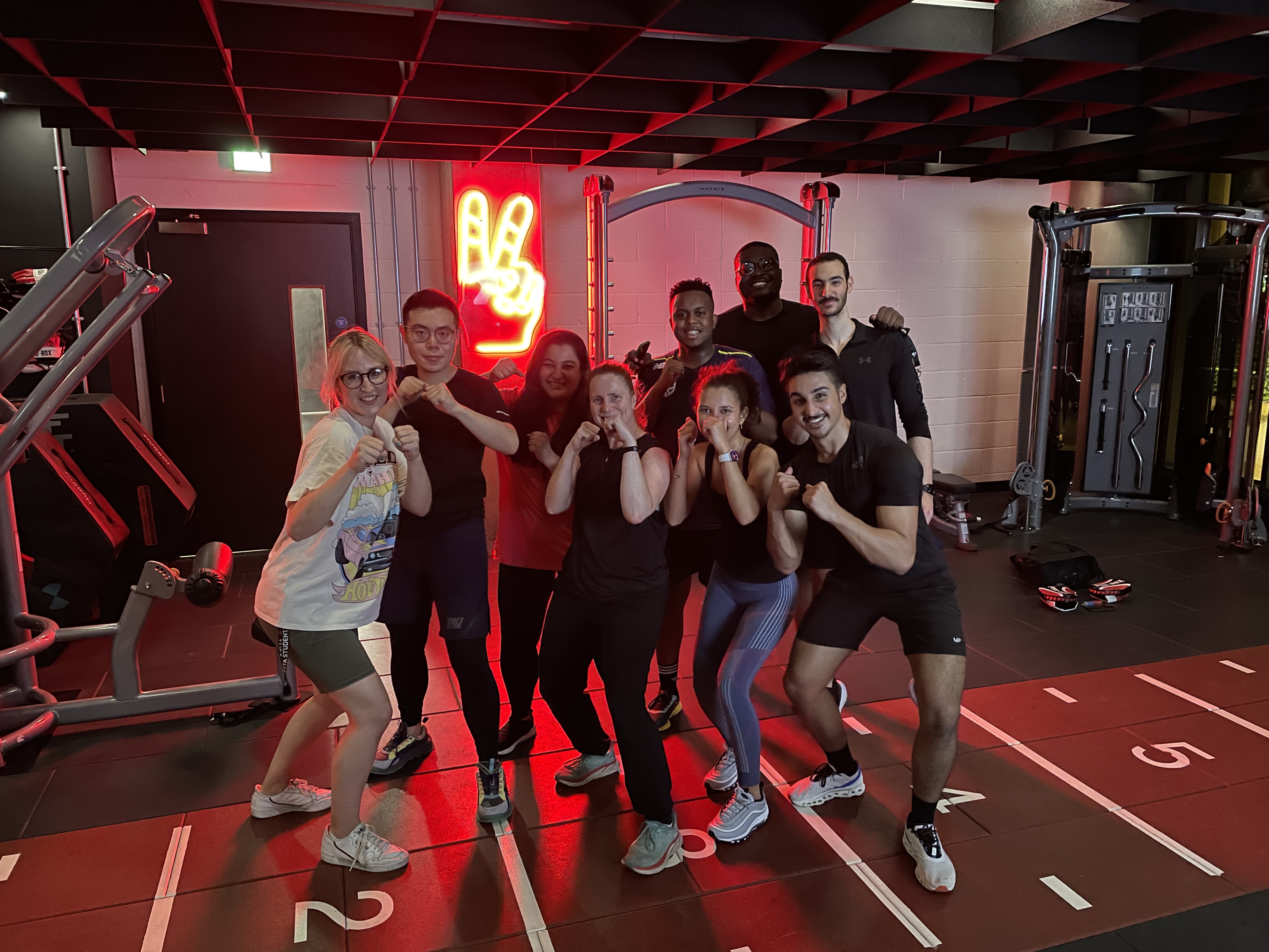 A group of eight people, dressed in workout clothes, stand in a gym posing with fists raised in a boxing stance. Behind them is a neon sign in the shape of a hand giving a peace sign. Exercise equipment is visible in the background.