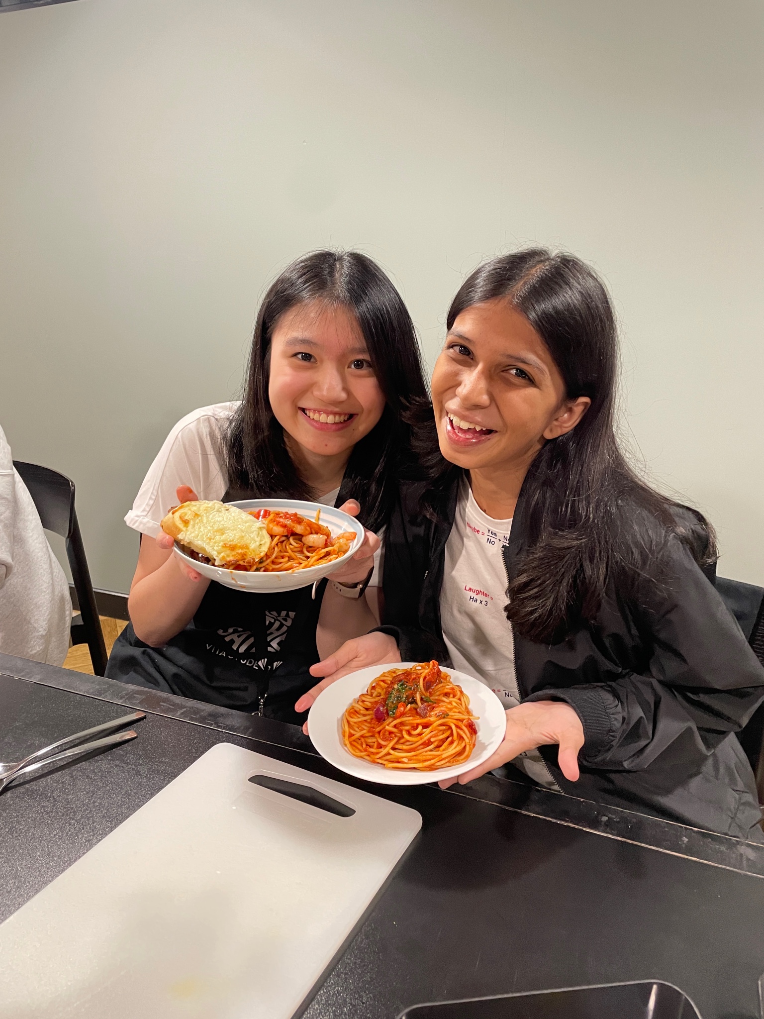 Two people smiling and holding plates of spaghetti. The person on the left has spaghetti with garlic bread, while the person on the right has spaghetti garnished with a herb. They are seated at a table with a white cutting board in front of them.