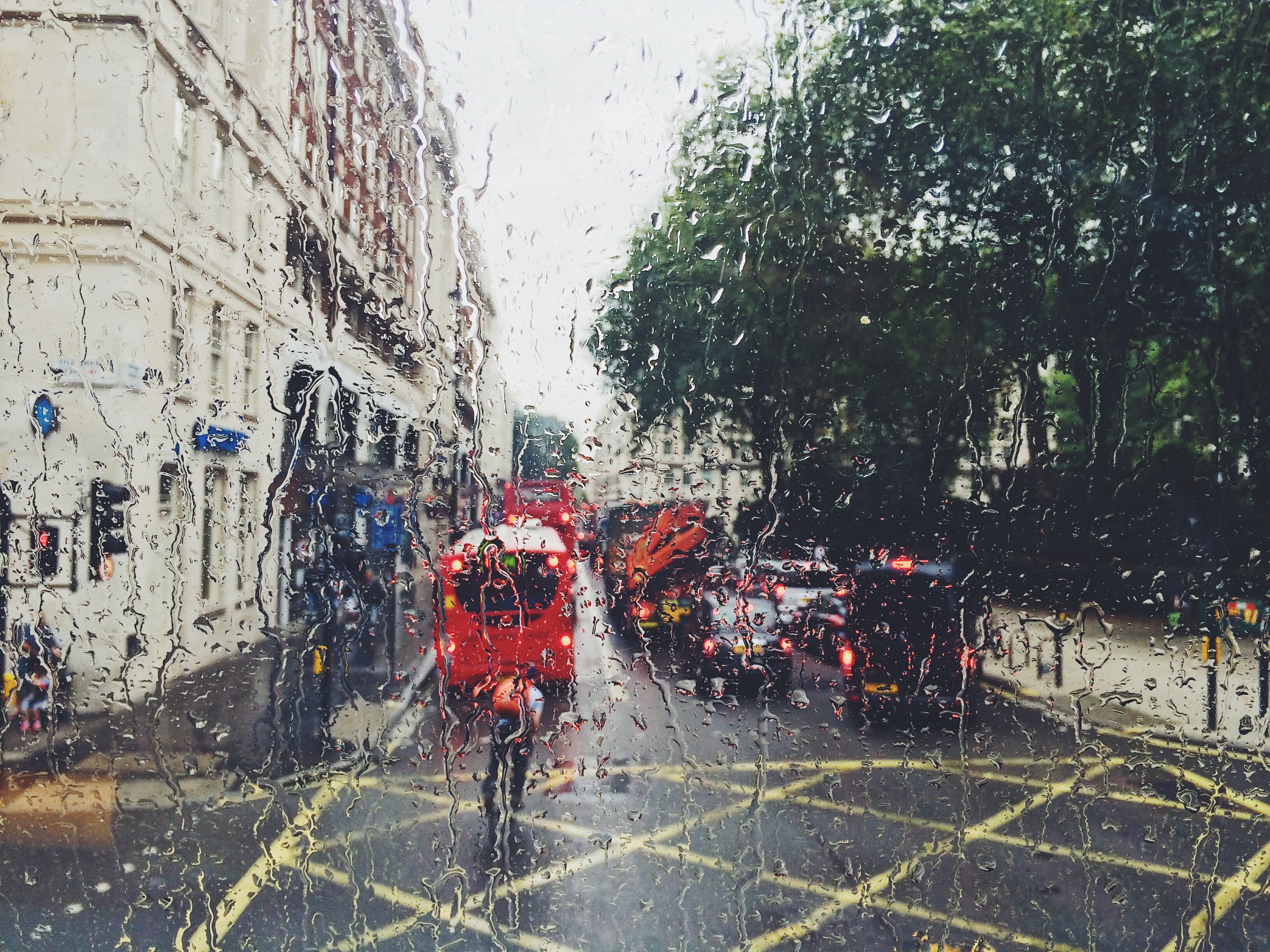 A rainy street scene unfolds through a window speckled with raindrops. Red double-decker buses and cars navigate the slick road. Tall buildings line the left, while green trees stand on the right—a quintessential British winter day in contrast to warm climates, teeming with life and culture.