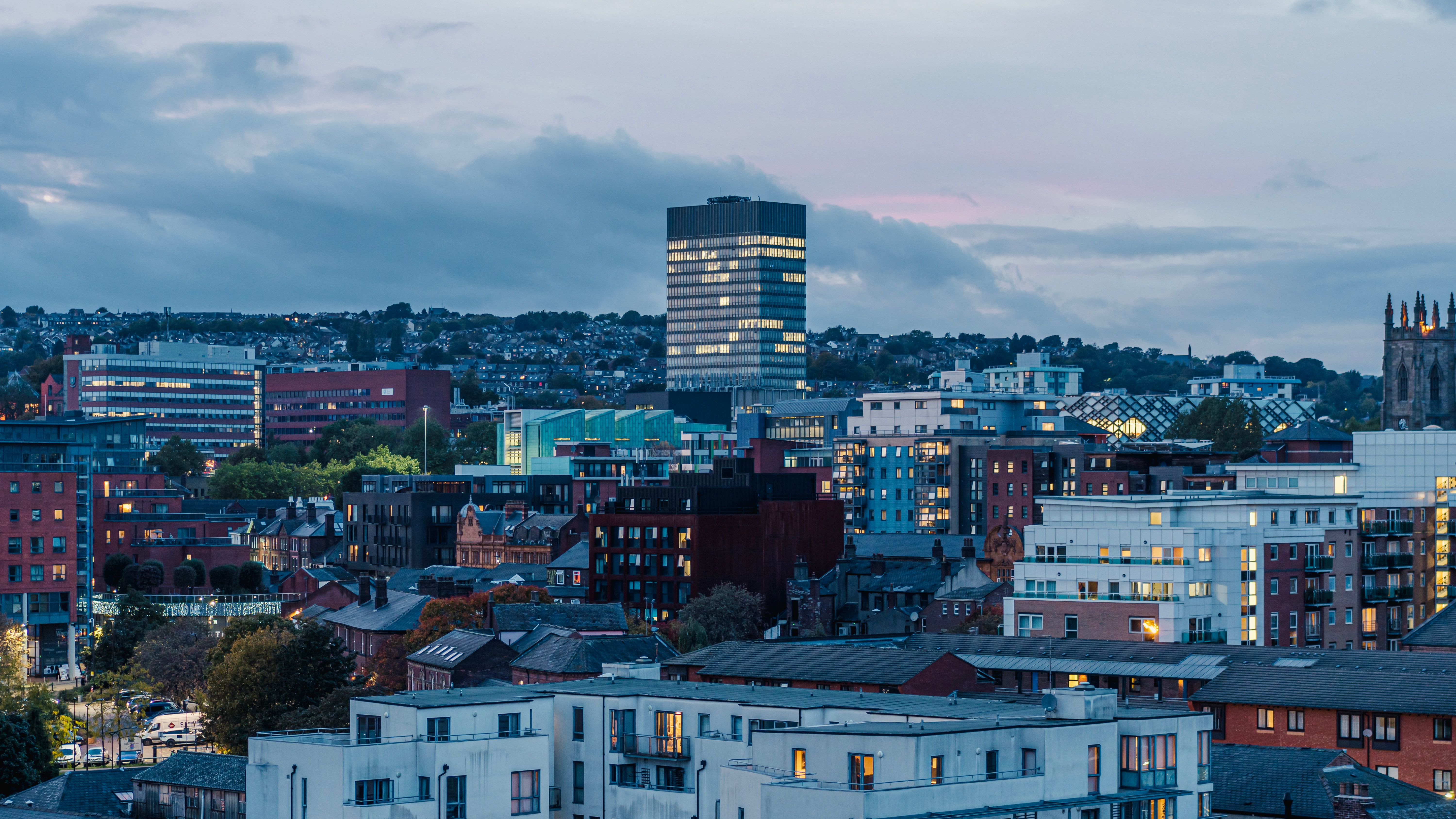 Cityscape at twilight featuring a mix of modern and traditional buildings in Sheffield. A tall, illuminated skyscraper stands prominently against the twilight sky. Rooftops in the foreground and a backdrop of rolling hills complete this captivating vista, perfect for exploring as part of any student guide.