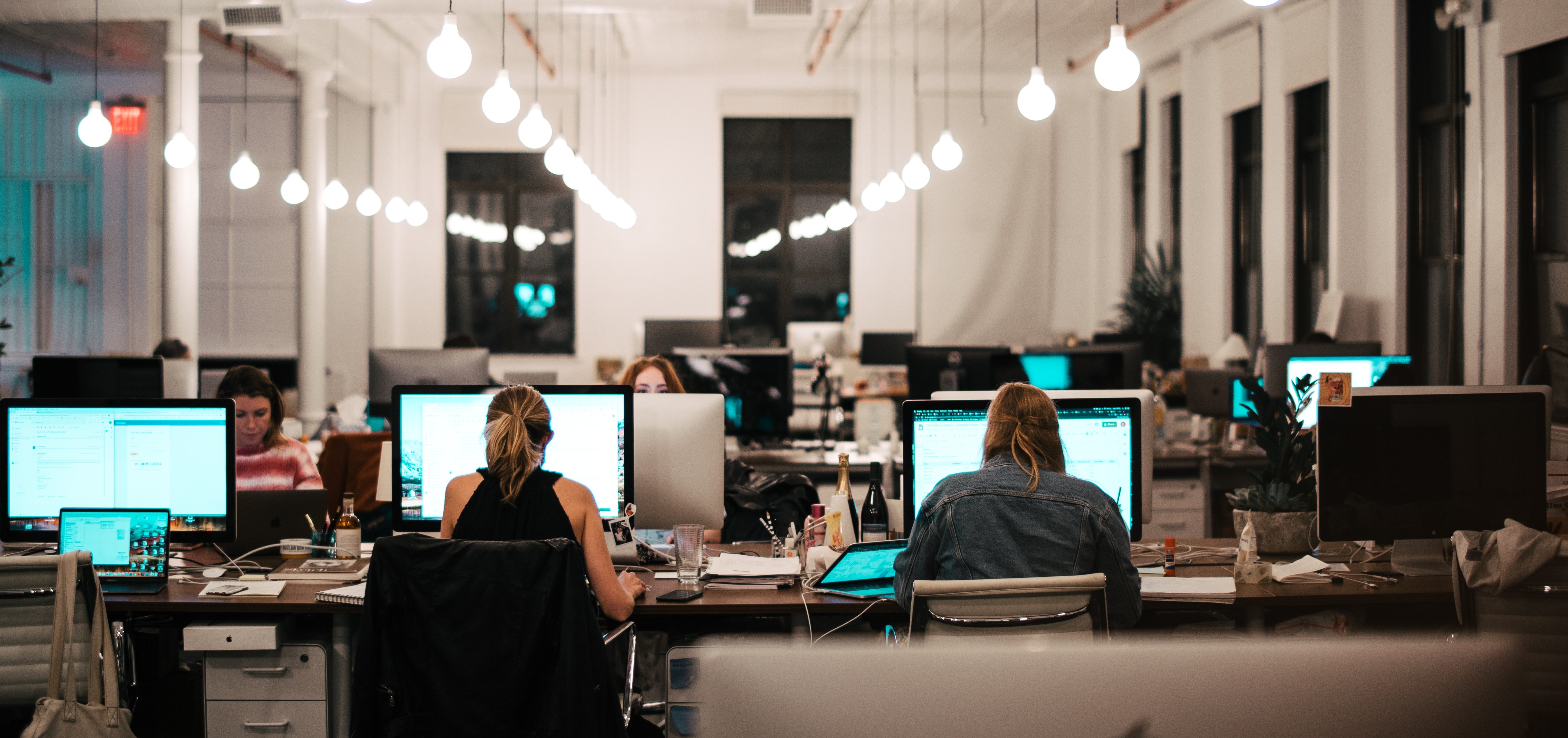 An open-plan office with several people working at desks, many discussing graduate schemes. Each desk has a computer monitor, and overhead lights illuminate the space. The environment appears focused and busy.