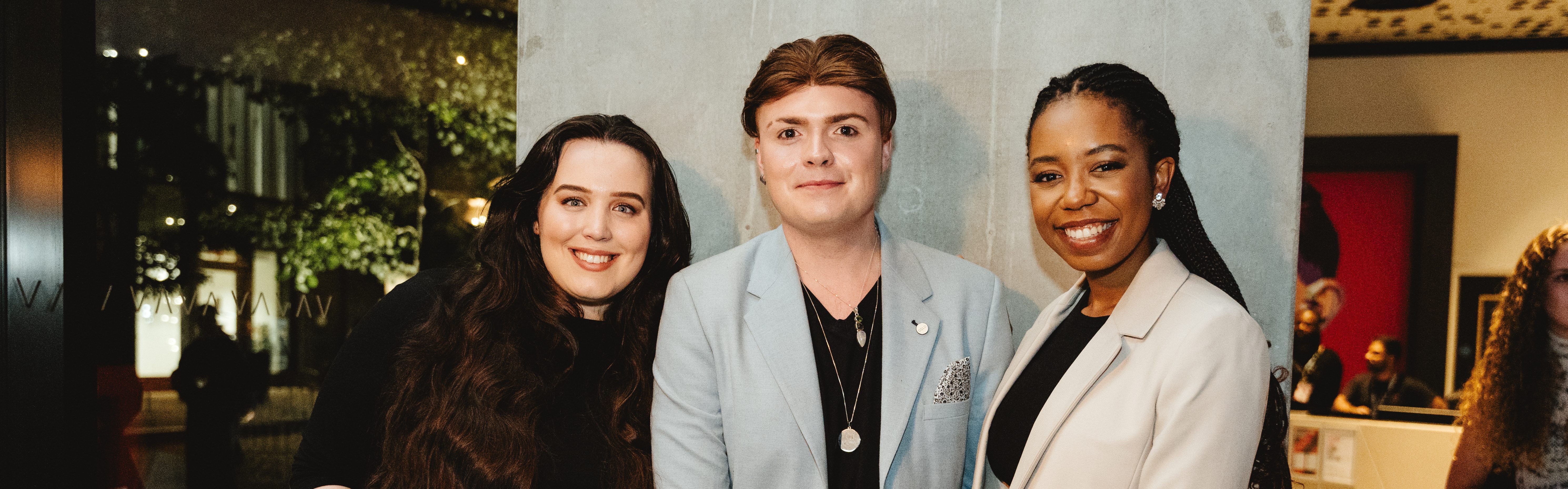Three people are smiling and posing together indoors, celebrating student careers. The person on the left has long dark hair, the person in the middle is wearing a light blue jacket, and the person on the right is in a cream-colored blazer. A blurred background shows artwork and greenery.