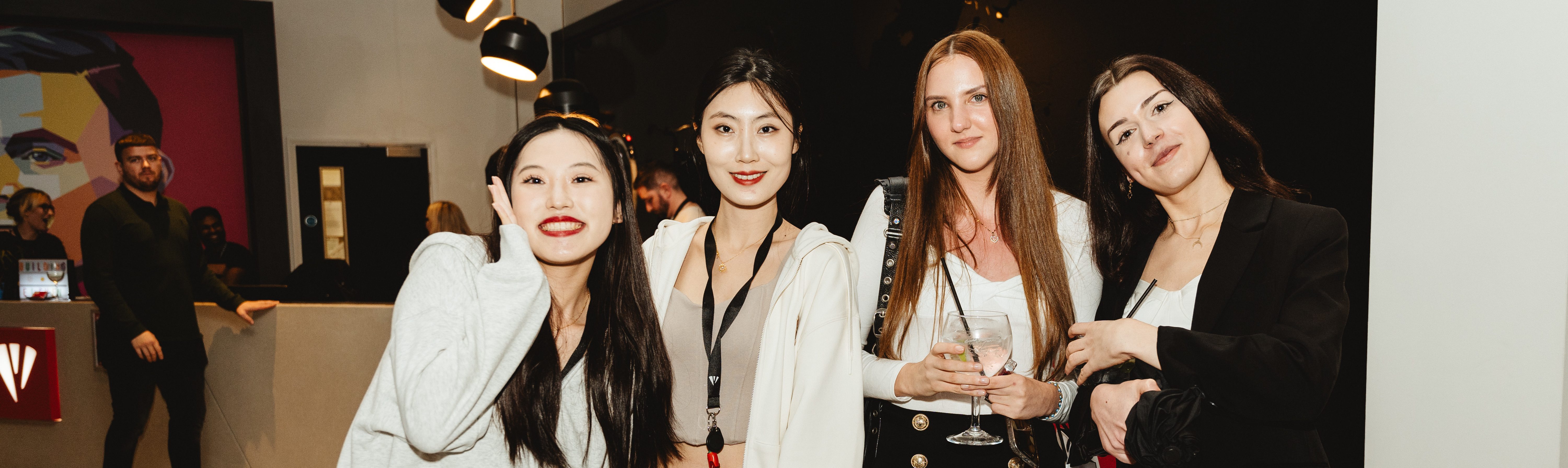 Four women are smiling and posing together in a brightly lit room, capturing the joy of a lively alumni reunion event. Three are wearing jackets, and one holds a glass. In the background, a mural and people add to the vibrant atmosphere at Vita Student.