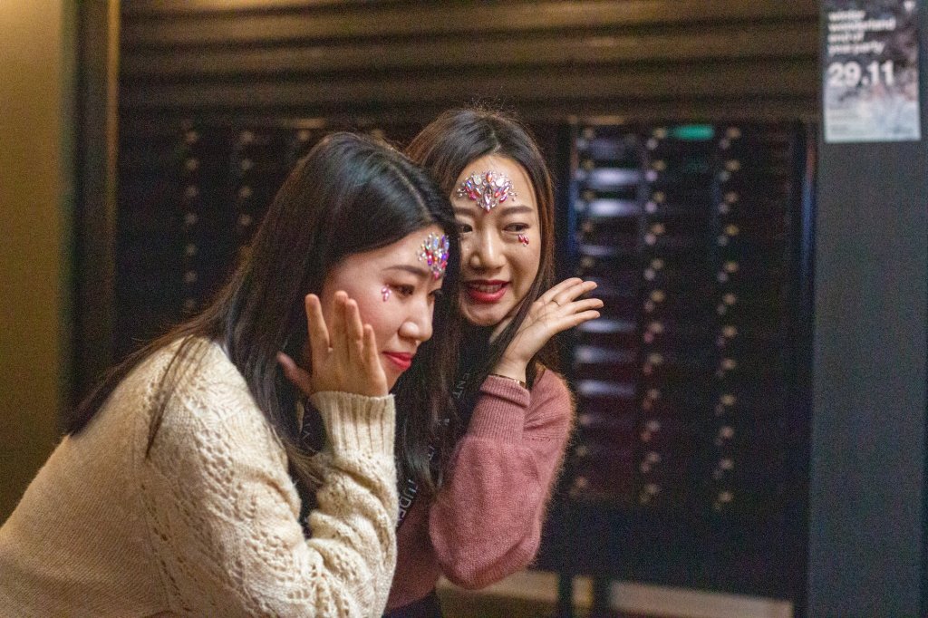 Two young women with face stickers and glitter smile and pose playfully together indoors in cozy sweaters—a perfect vibe for enjoying fun Birmingham activities or exploring things to do in Birmingham with friends. The dim background features dark panels and a blurry poster.