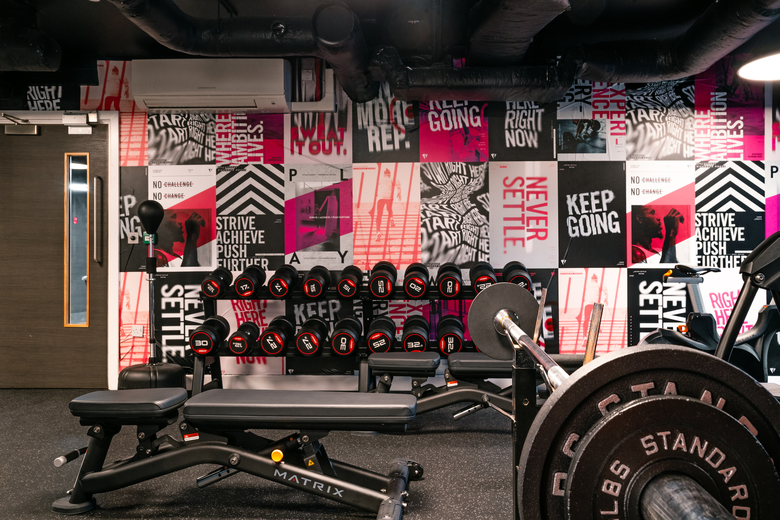 The gym room in the student accommodation features motivational wall art with phrases like "Keep Going" and "Never Settle." Equipped with benches, dumbbells, barbells, and diverse weightlifting gear, the floor is covered with black rubber mats for a safe workout experience.