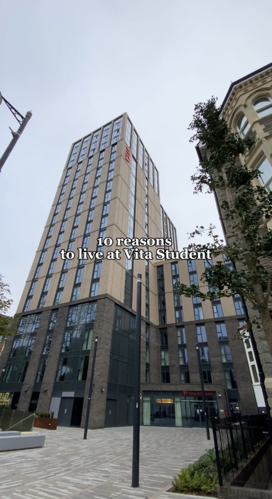 A tall, modern apartment building with large windows viewed from the street. Text overlay reads "10 reasons to live at Vita Student." Trees and other buildings are partially visible in the foreground.