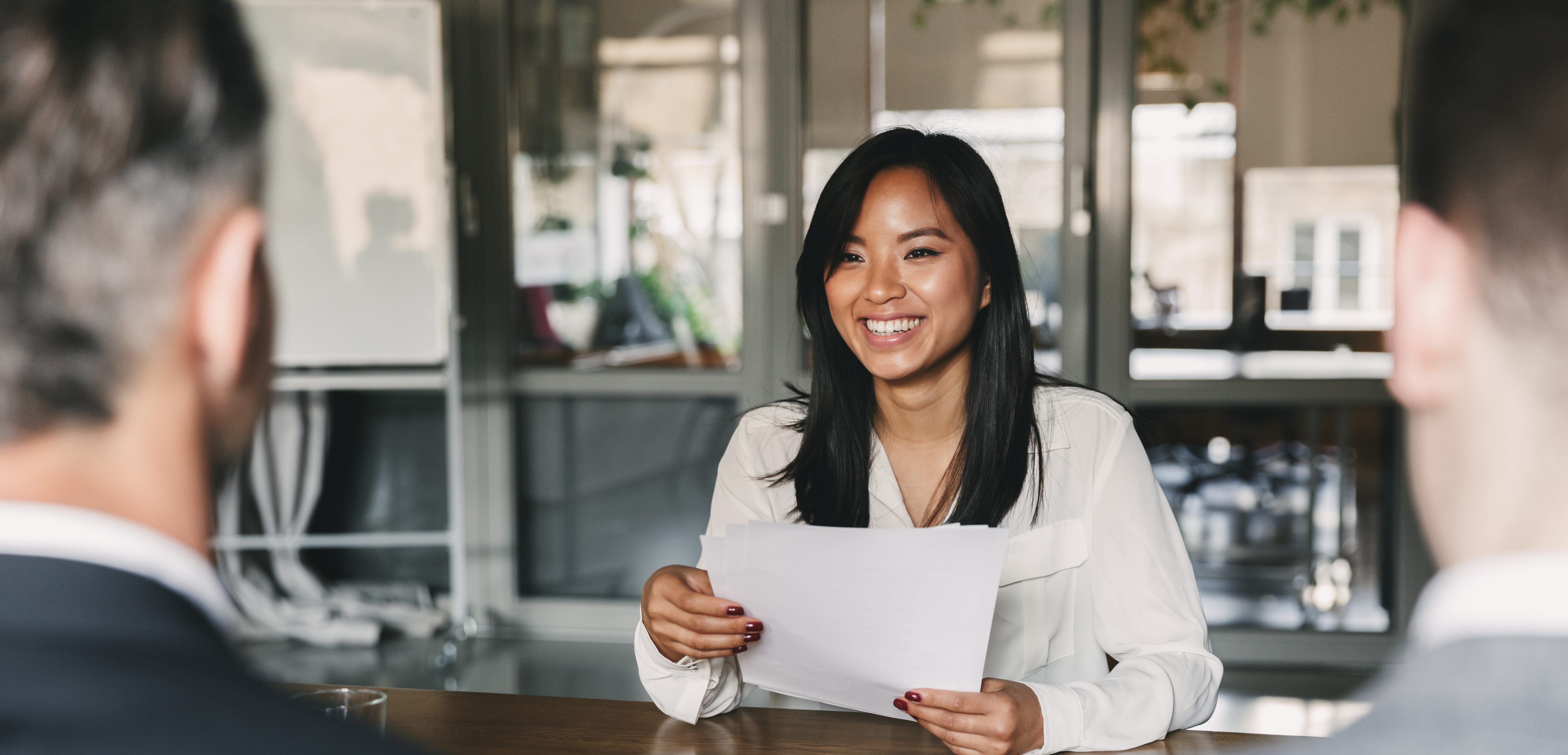 A woman with long black hair, wearing a white blouse, is smiling while holding papers. She sits across a table from two people in a bright office setting, possibly sharing tips during an interview or meeting.