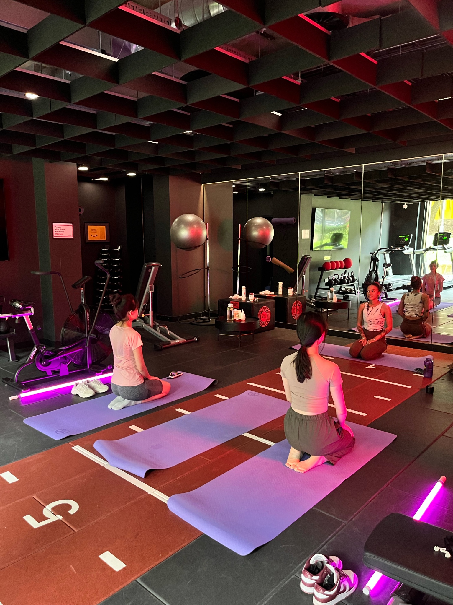 Three women in a gym are sitting on yoga mats, facing a mirrored wall. The room has dim lighting with pink accents. University branding is subtly woven into the decor, while exercise equipment like weights and fitness balls is visible in the background.