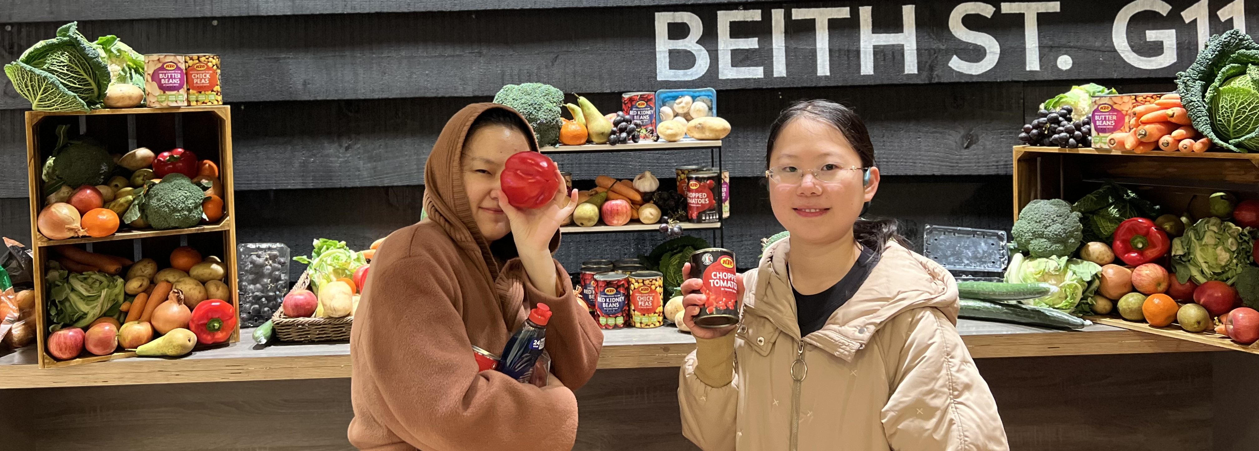 Two students stand smiling in front of a vibrant vegetable display with produce like peppers and broccoli. One playfully holds a red bell pepper over an eye, while the other clutches a can of coconut sugar. Behind them, a Beith Street sign adds charm to this Veganuary-inspired scene.