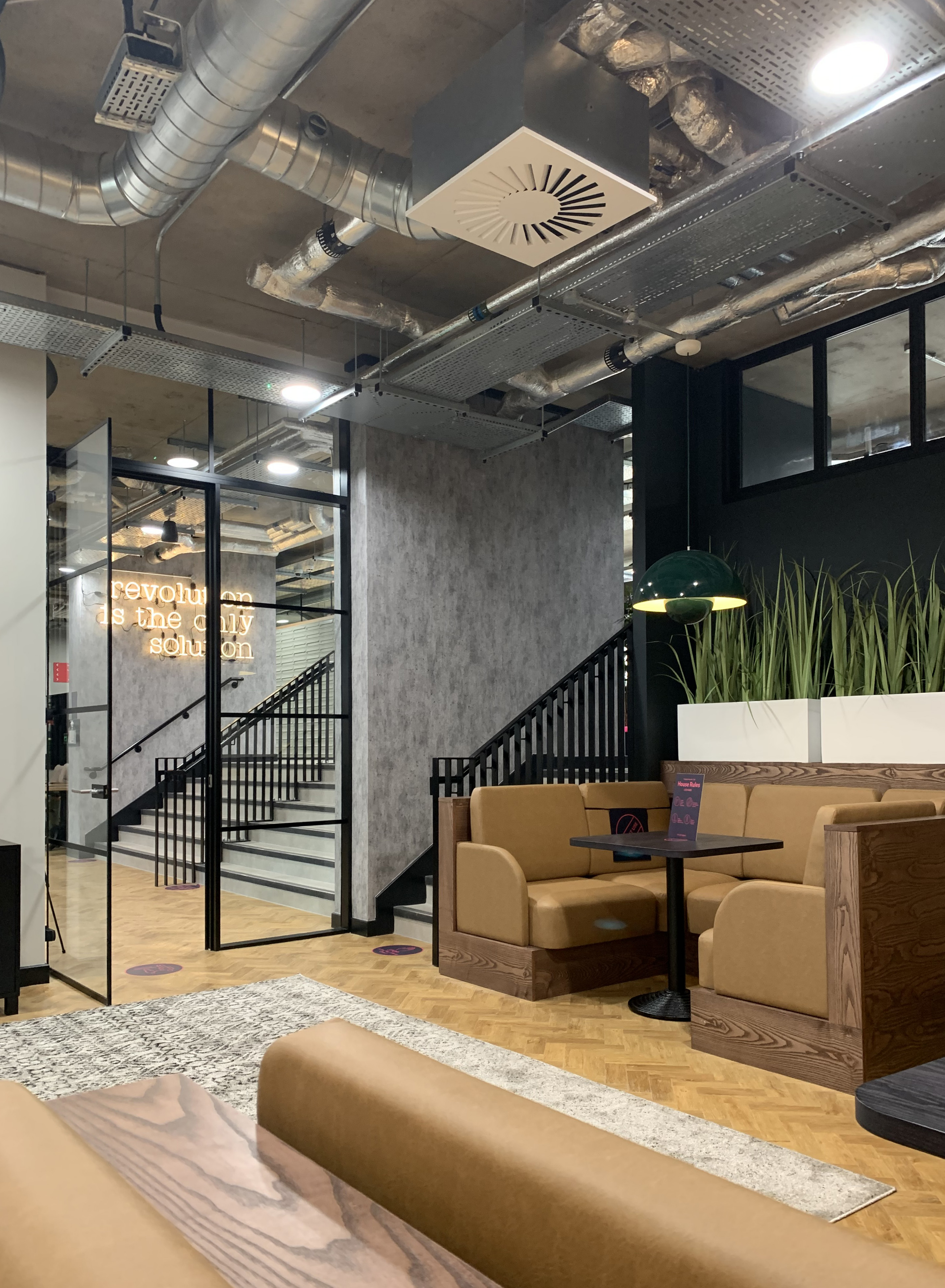 Modern office space with exposed ductwork and industrial design. Features include brown leather seating, a wooden coffee table, wall planters, a green hanging lamp, and a sign reading "revolution is the only solution" near the stairs.