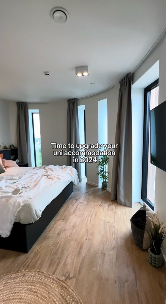 Modern bedroom with a large bed, white bedding, and tall windows. Light wood flooring and grey curtains add to the decor. A wall-mounted TV and a small plant are present. Text reads: "Time to upgrade your uni accommodation in 2024.