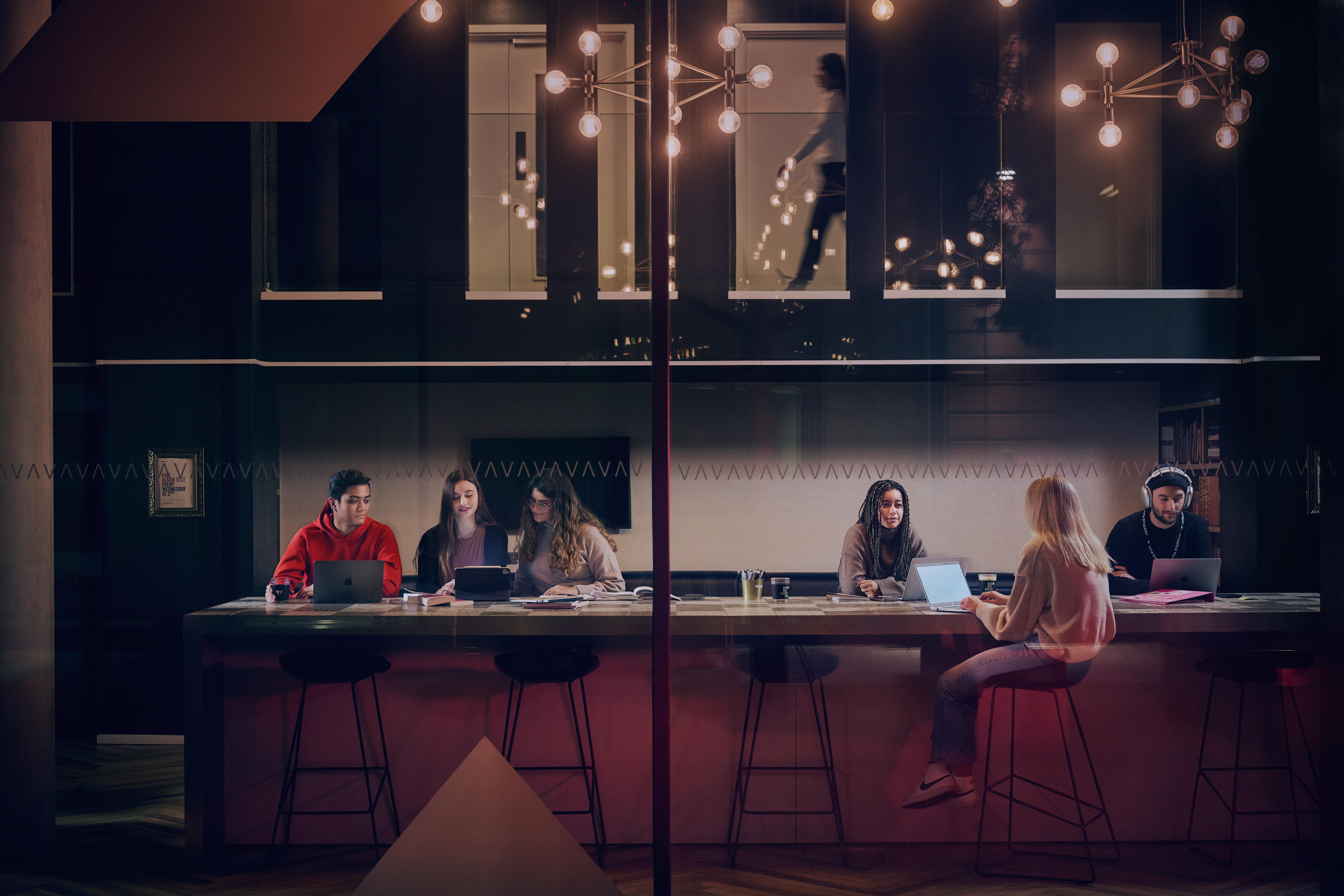 A group of people work at a long table with laptops and papers in a modern room with dim lighting. Sleek chandeliers hang from the ceiling, casting a warm glow. Through the glass wall, a blurred figure walks by outside, set against the urban backdrop of Newcastle's bustling city streets.